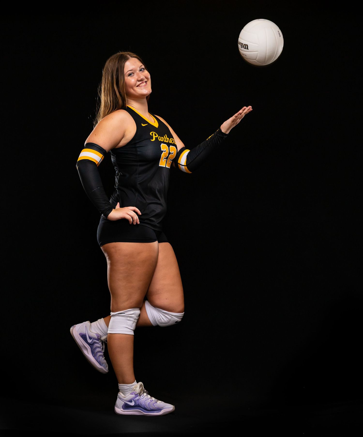Smiling female Panthers volleyball player #22 tossing a Wilson volleyball in black and gold uniform.