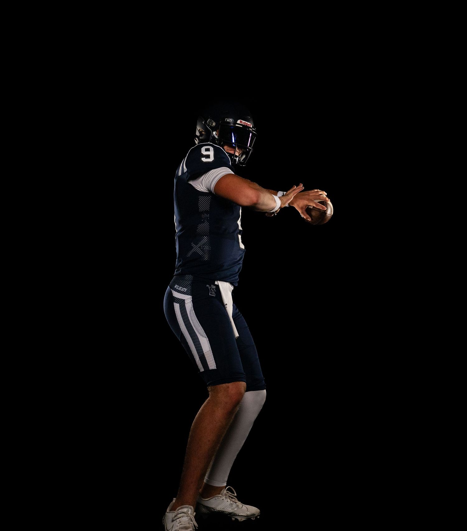 Football player #9 in navy uniform in throwing stance against black background.