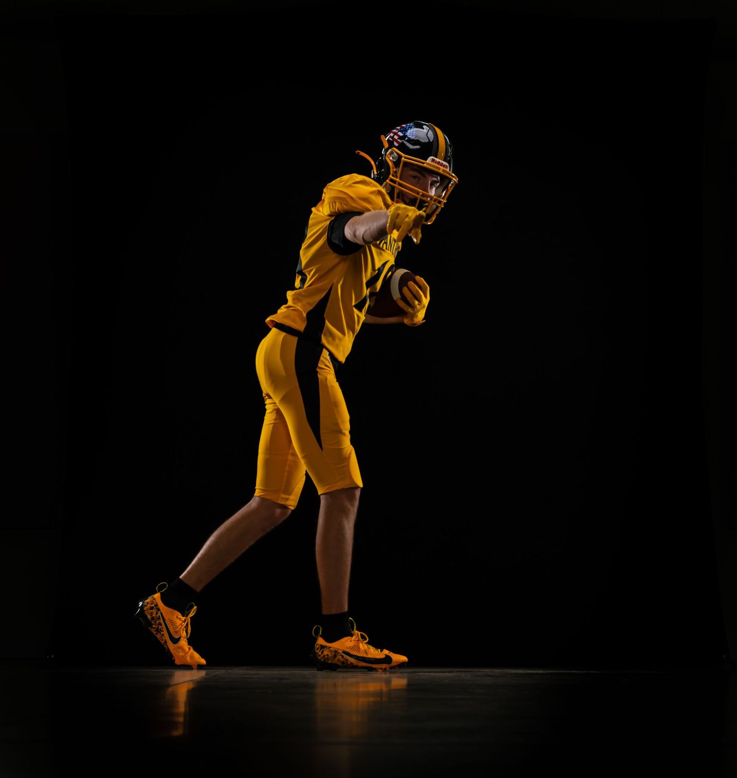 A football player in a gold uniform and helmet holds a ball in a dramatic dark studio setting.