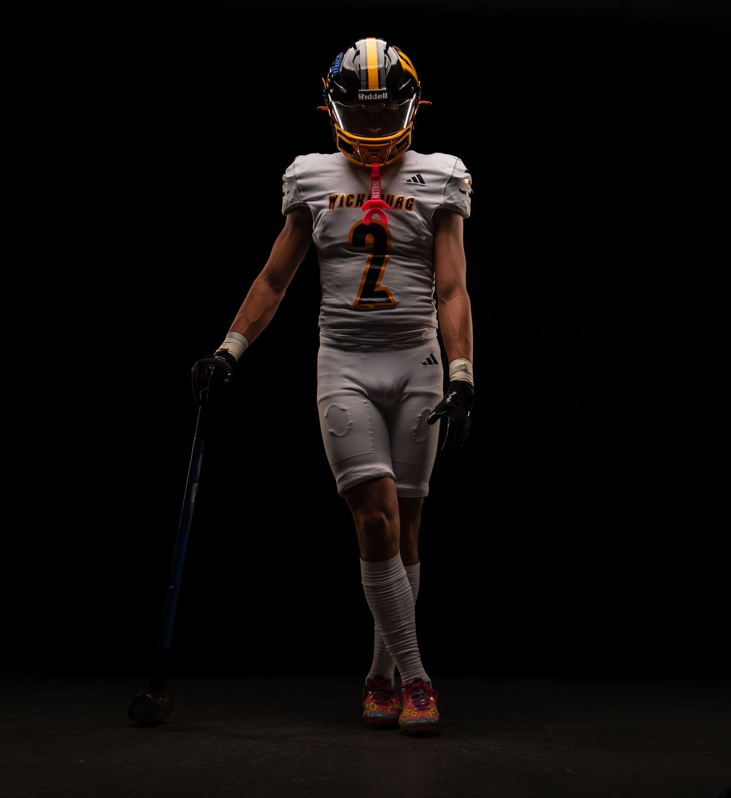 Football player in gray uniform with orange helmet stands dramatically against dark background.