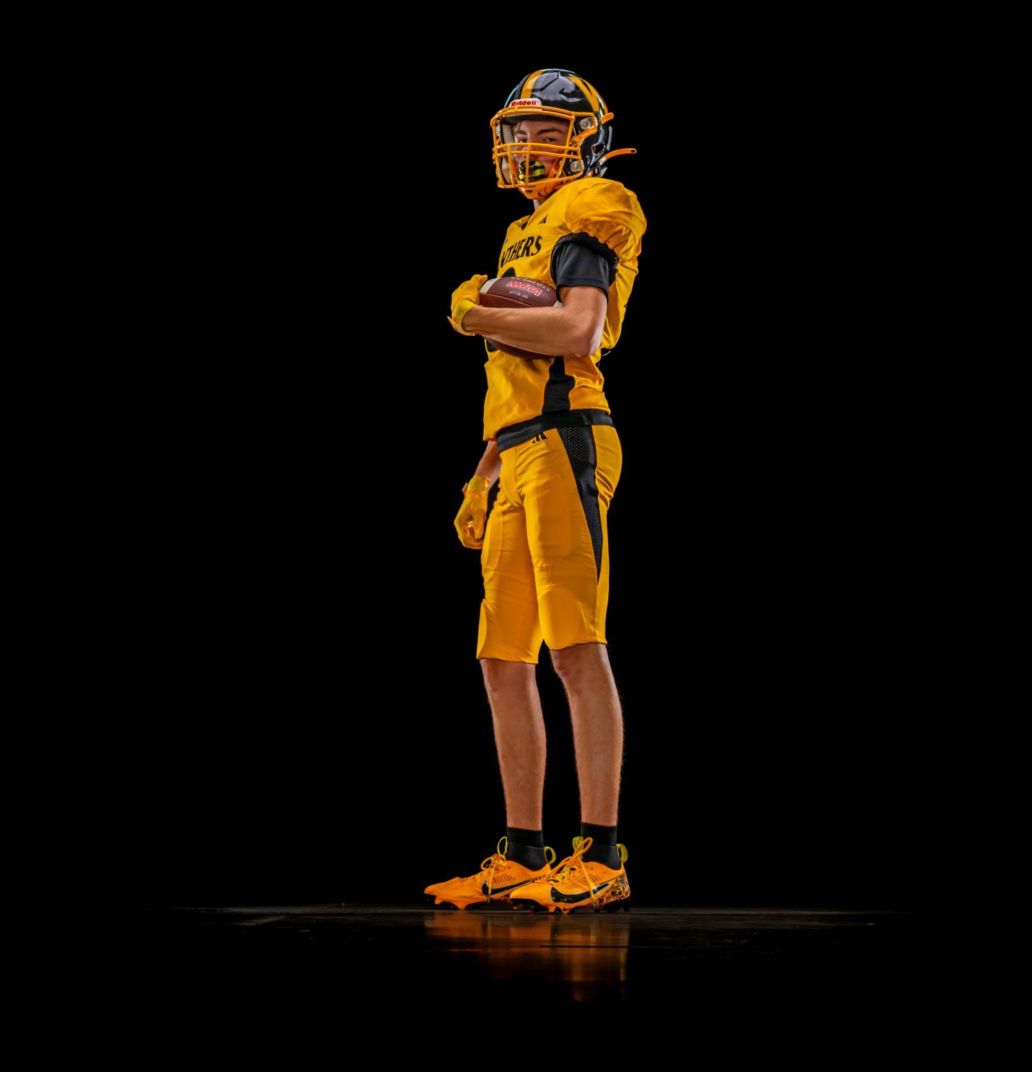 Young football player in gold uniform standing confidently with arms crossed on dark stage.