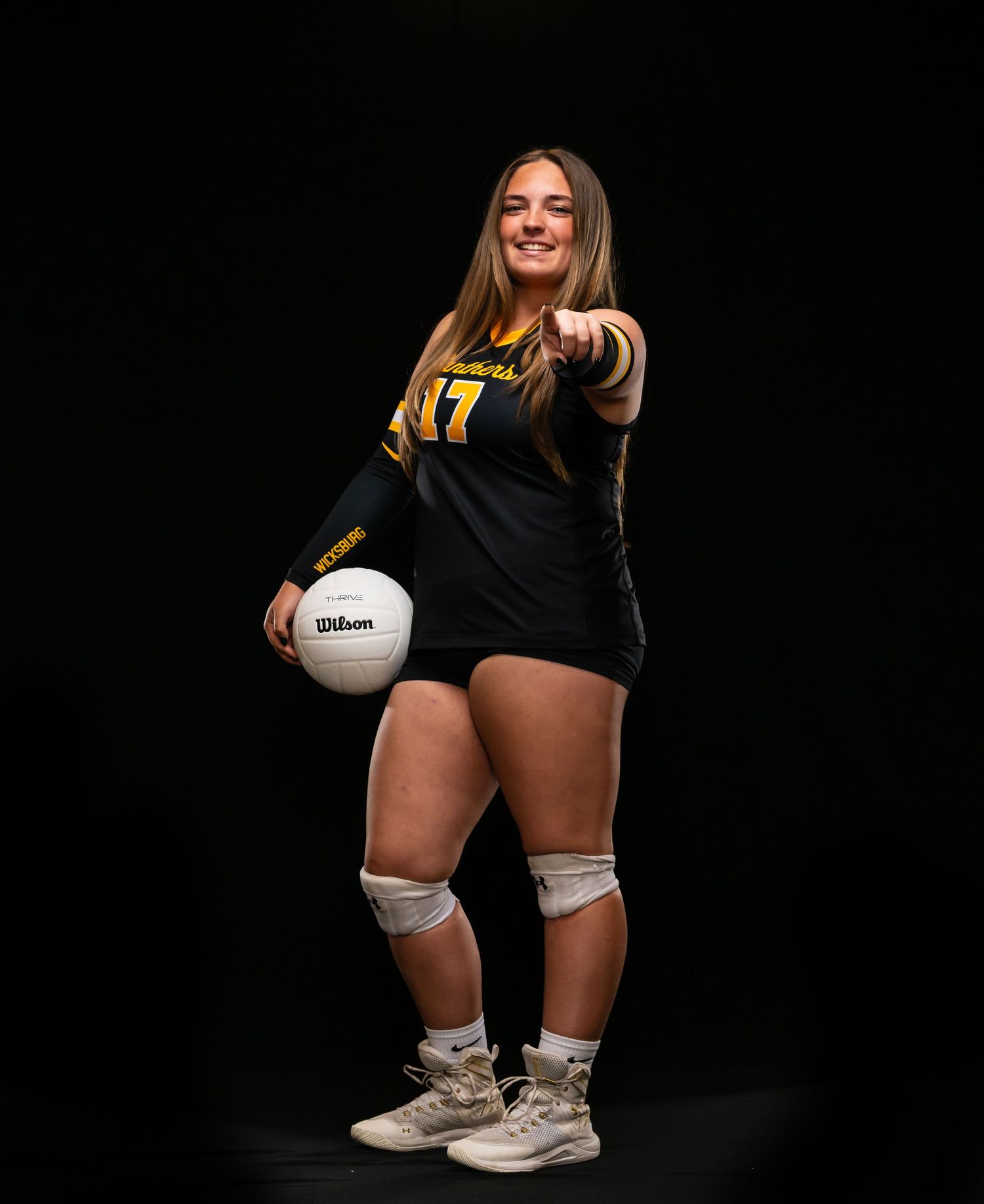 Smiling female volleyball player in black and gold jersey number 17, holding Wilson ball, pointing at camera.