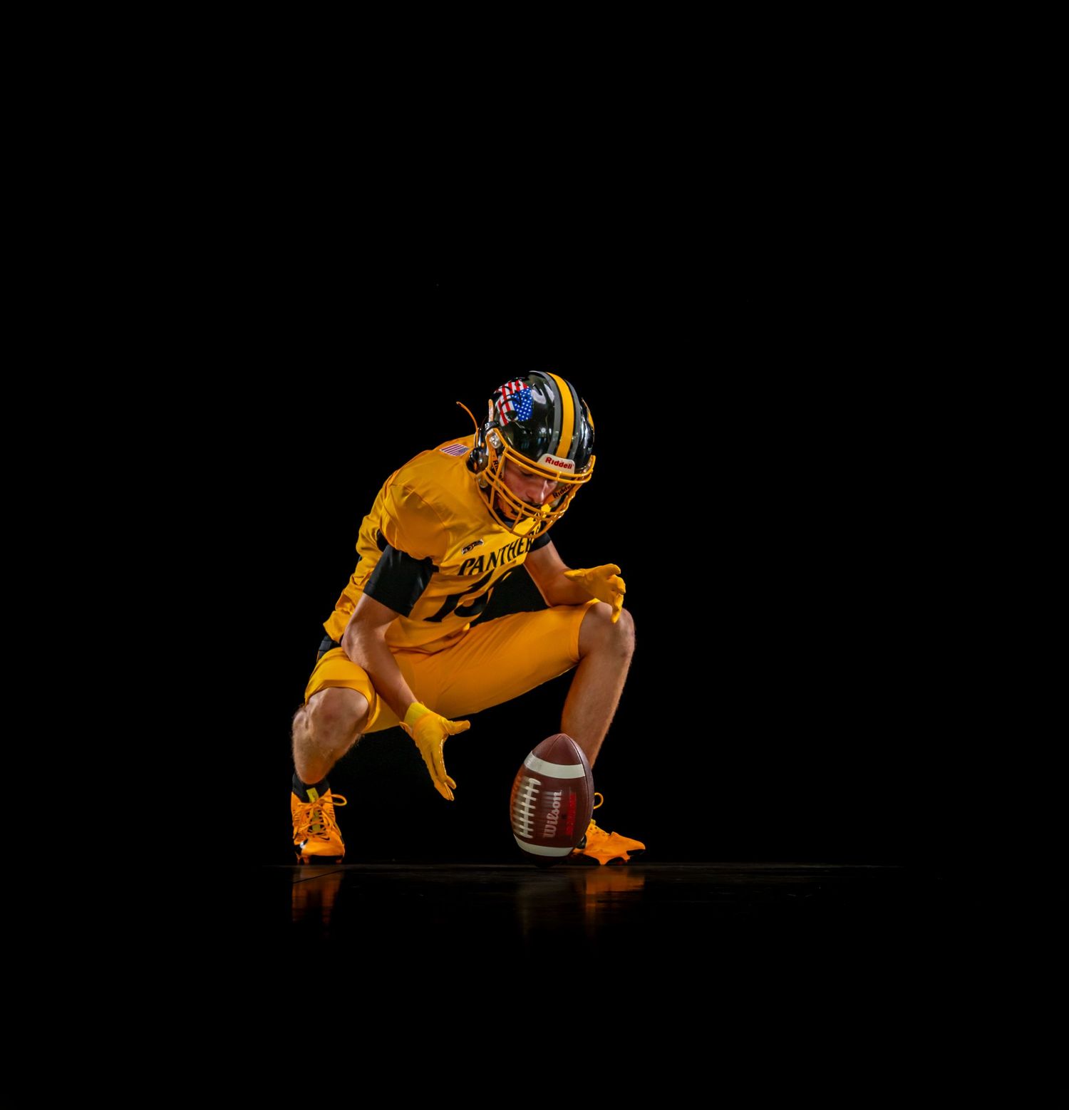 Football player in gold uniform crouches with ball on dark studio background.