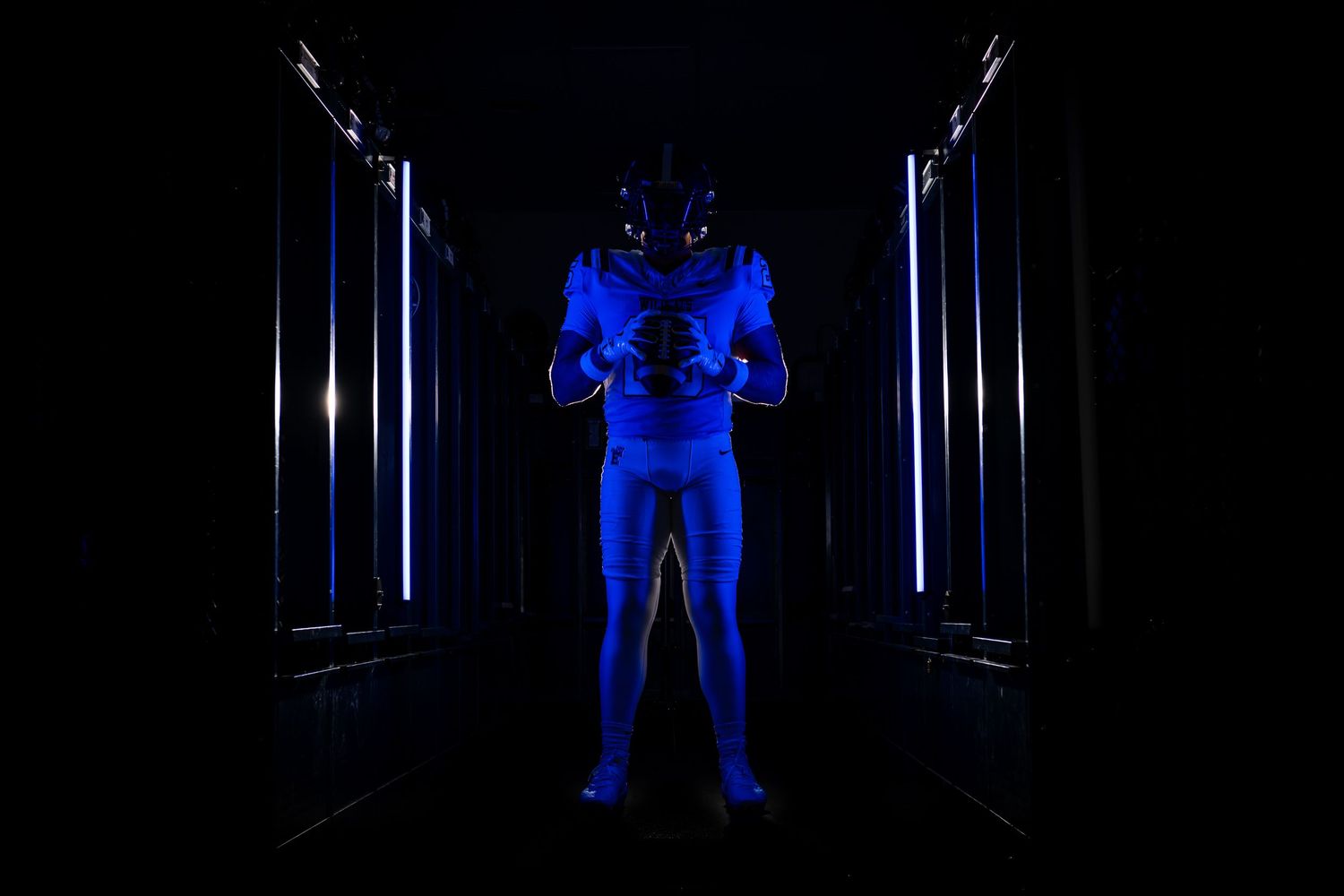 Football player in blue uniform and helmet illuminated by blue neon lights in dark corridor.