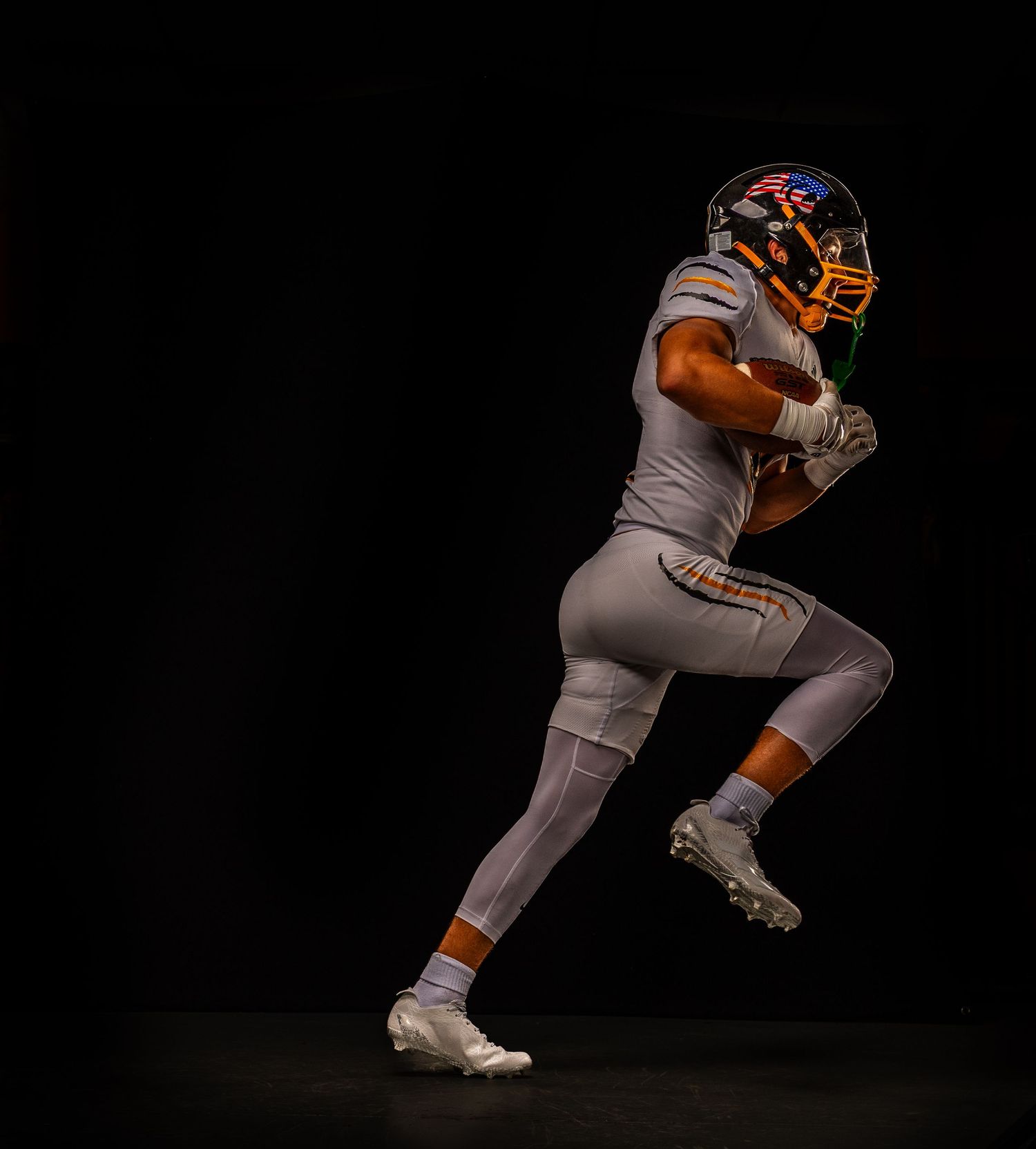 Football player in gray and orange uniform mid-stride against black background, wearing orange helmet and white cleats.