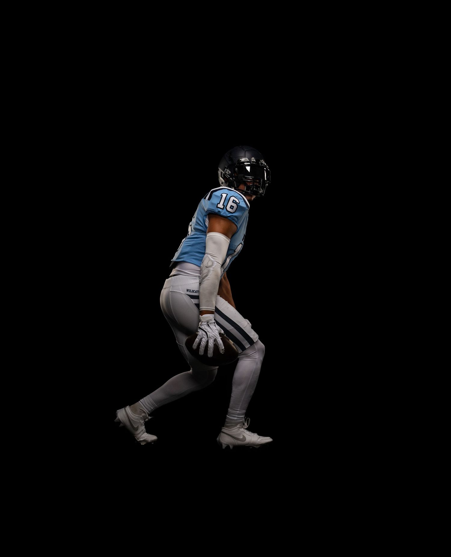 Football player #16 in blue jersey in a defensive stance against a black background.