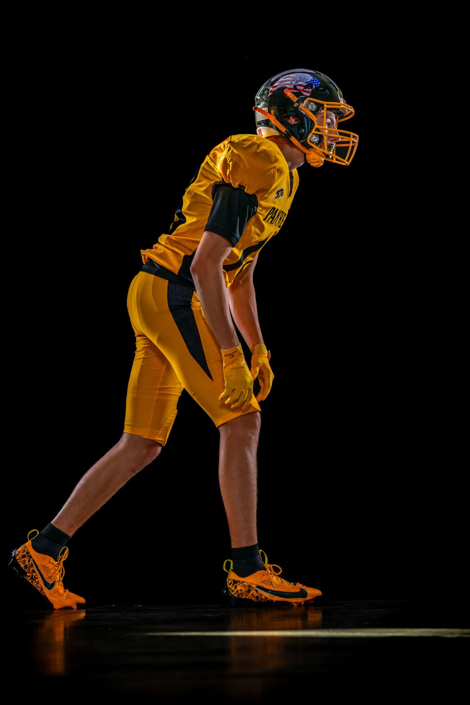 Young football player in gold uniform and helmet posed in athletic stance against black background.