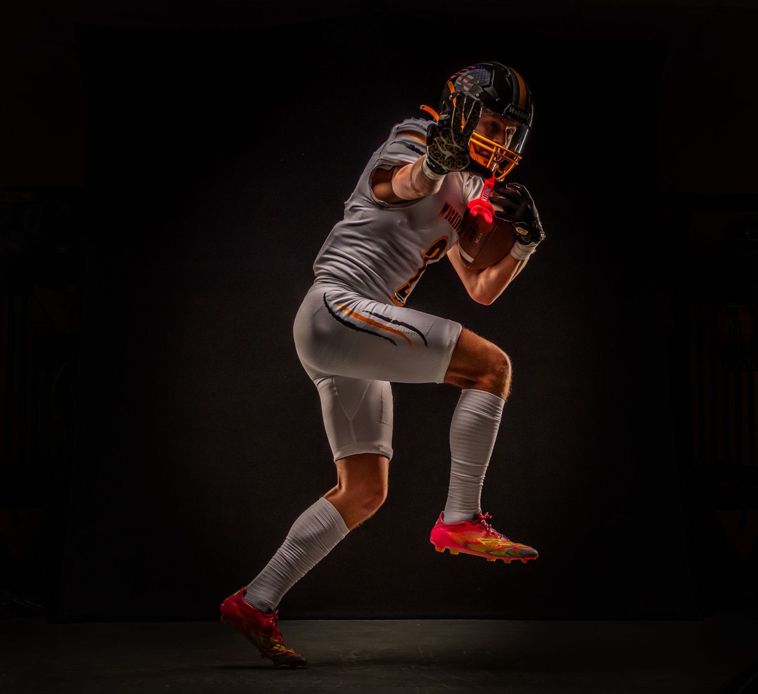 Football player in grey uniform strikes dynamic pose against dark background with dramatic lighting.