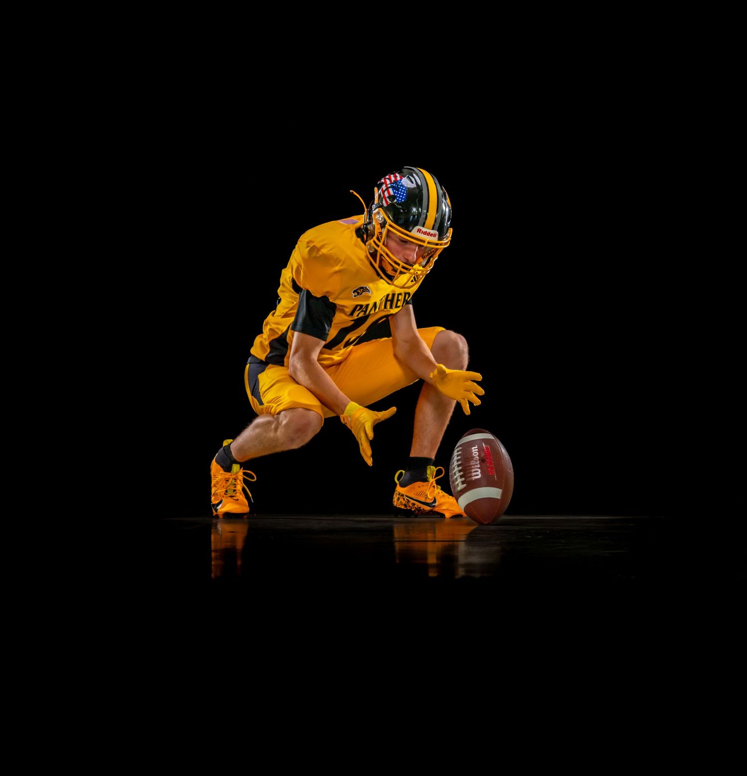 Youth football player in yellow uniform crouching to pick up a football against a dark background.