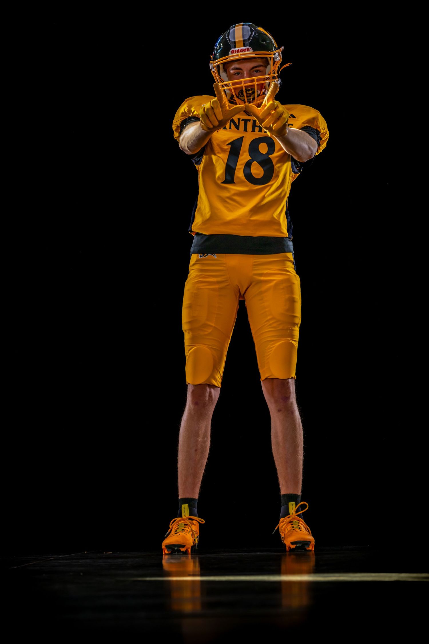 American football player #18 poses in gold uniform with hands raised on dark studio background.
