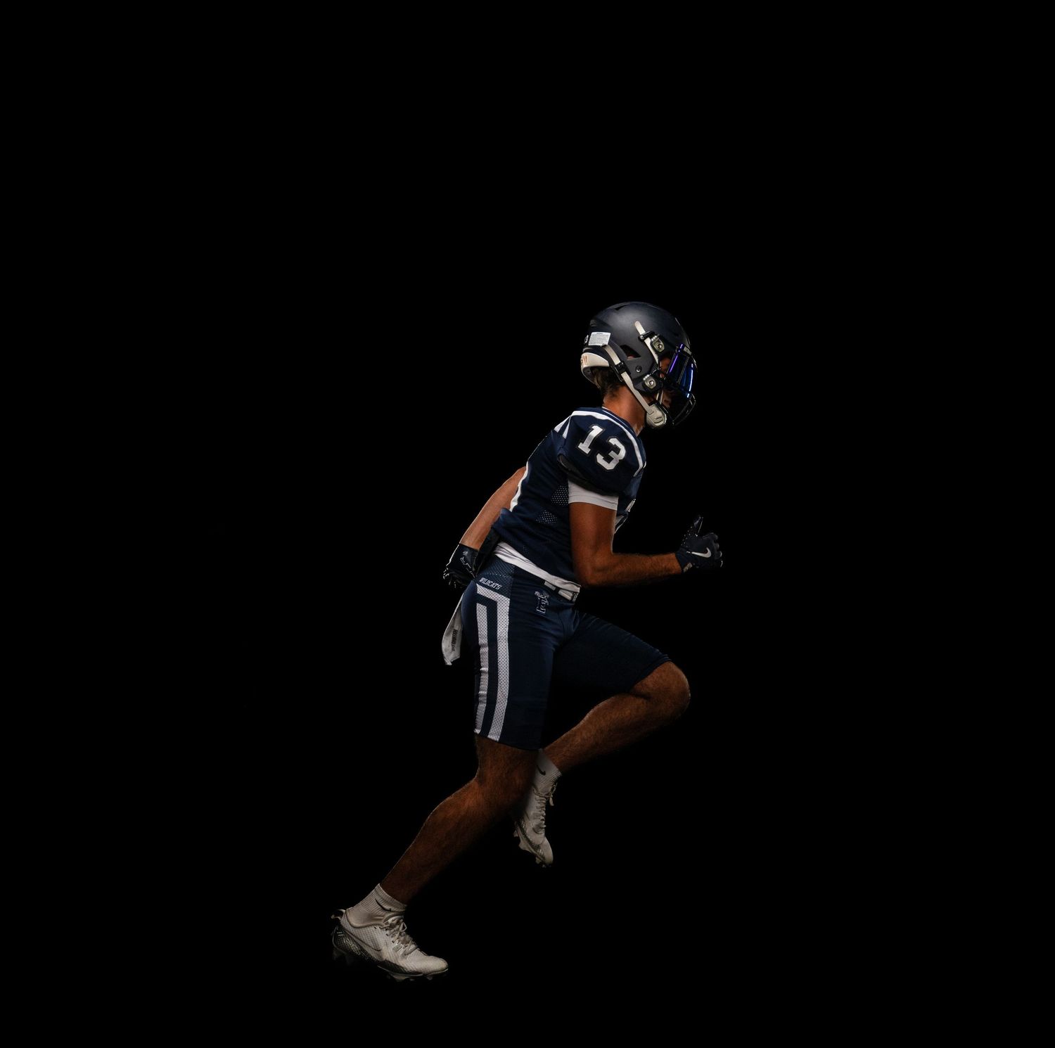 Football player number 13 in navy uniform sprints against dark background.