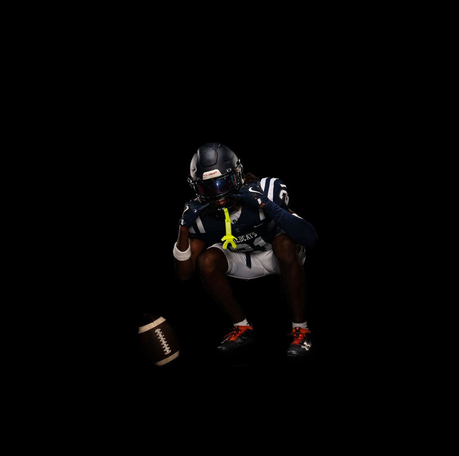 Football player in navy uniform crouching on dark background with football on ground beside them.