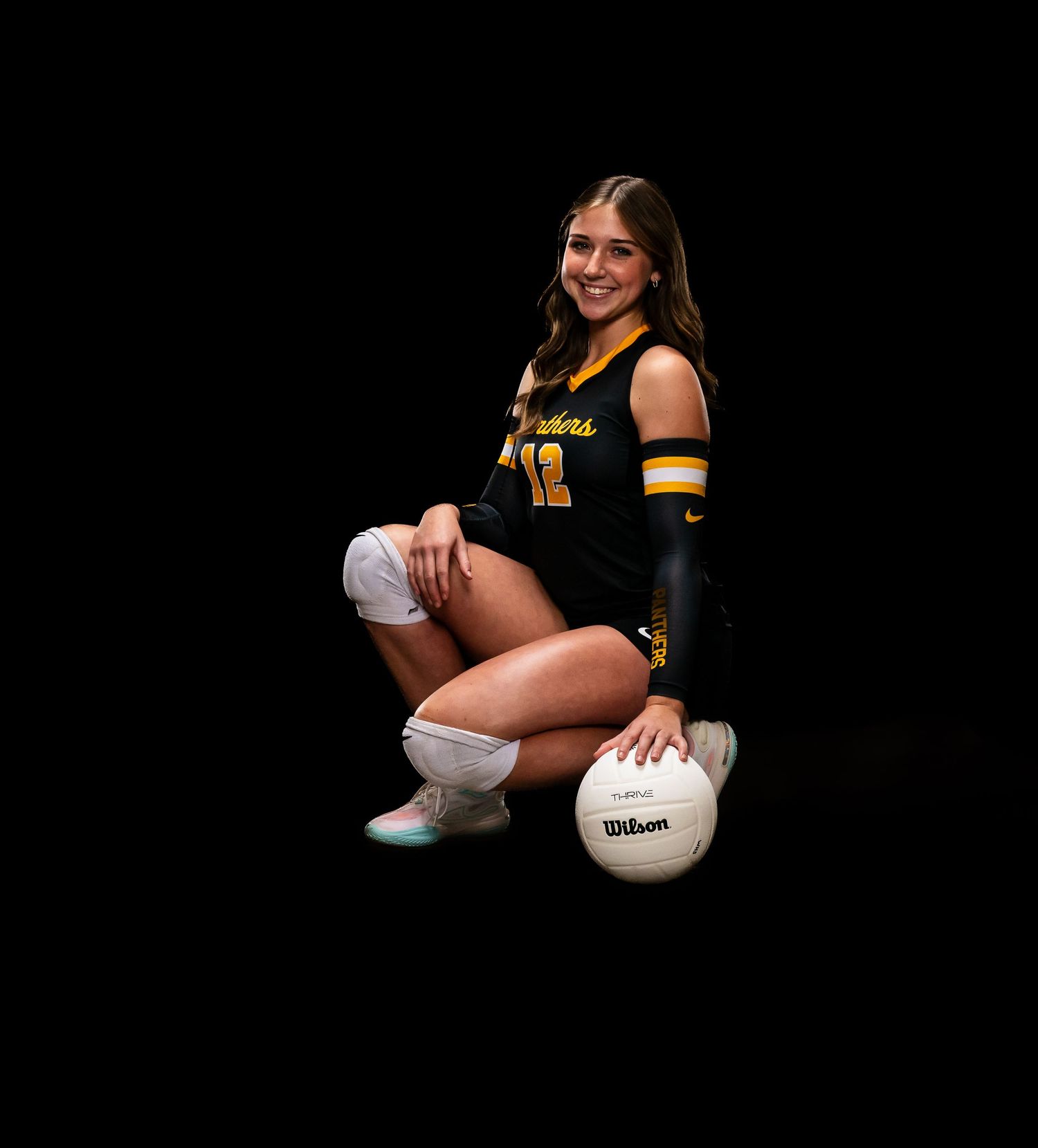 Female volleyball player in black and gold uniform posing with Wilson volleyball in professional sports portrait.