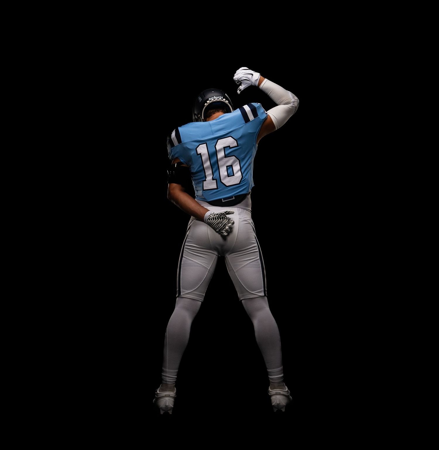 Football player #16 in blue jersey flexing with arms raised against a black background.