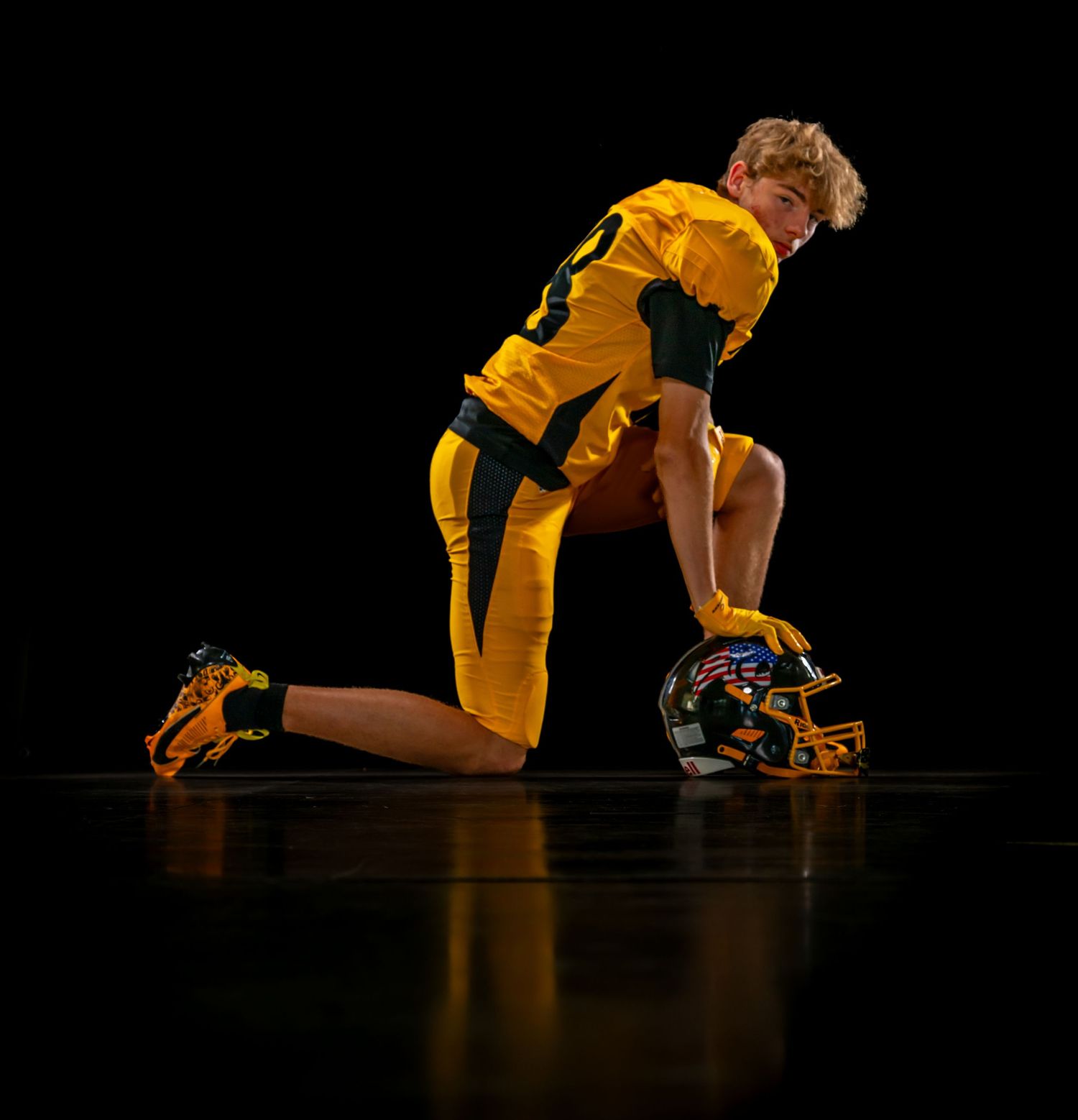 Football player in gold uniform kneels on one knee holding helmet on dark background.