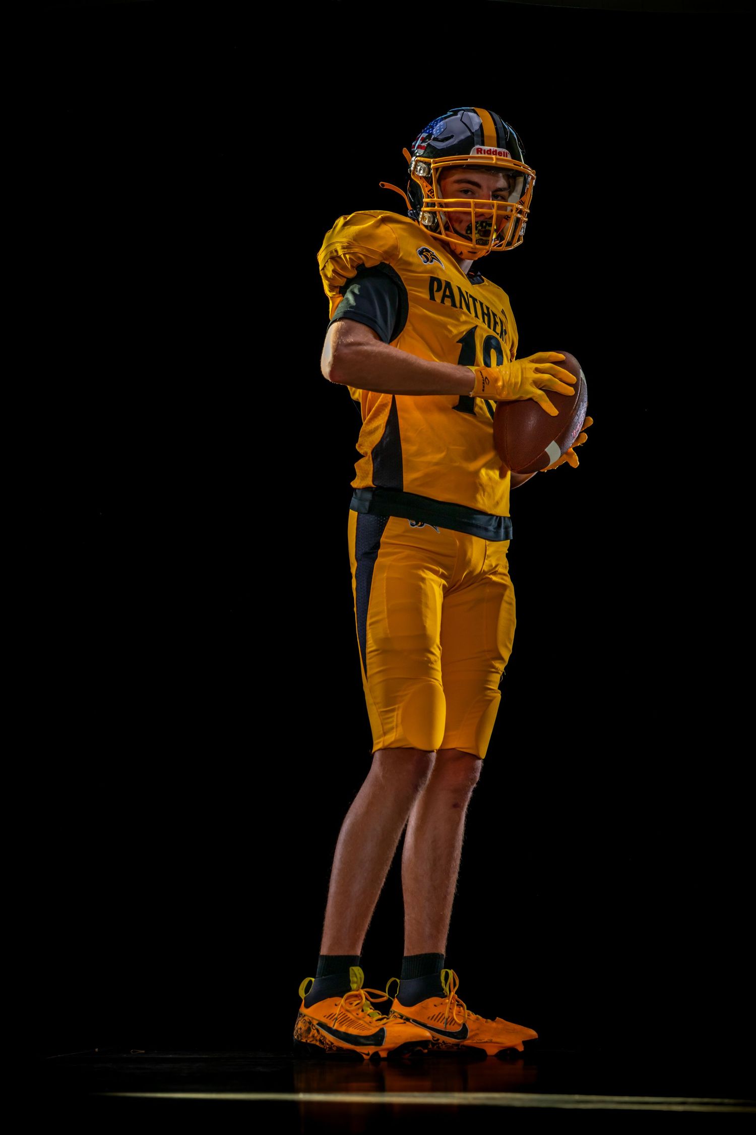 Young football player in yellow Panthers uniform and helmet holds football against dark background.