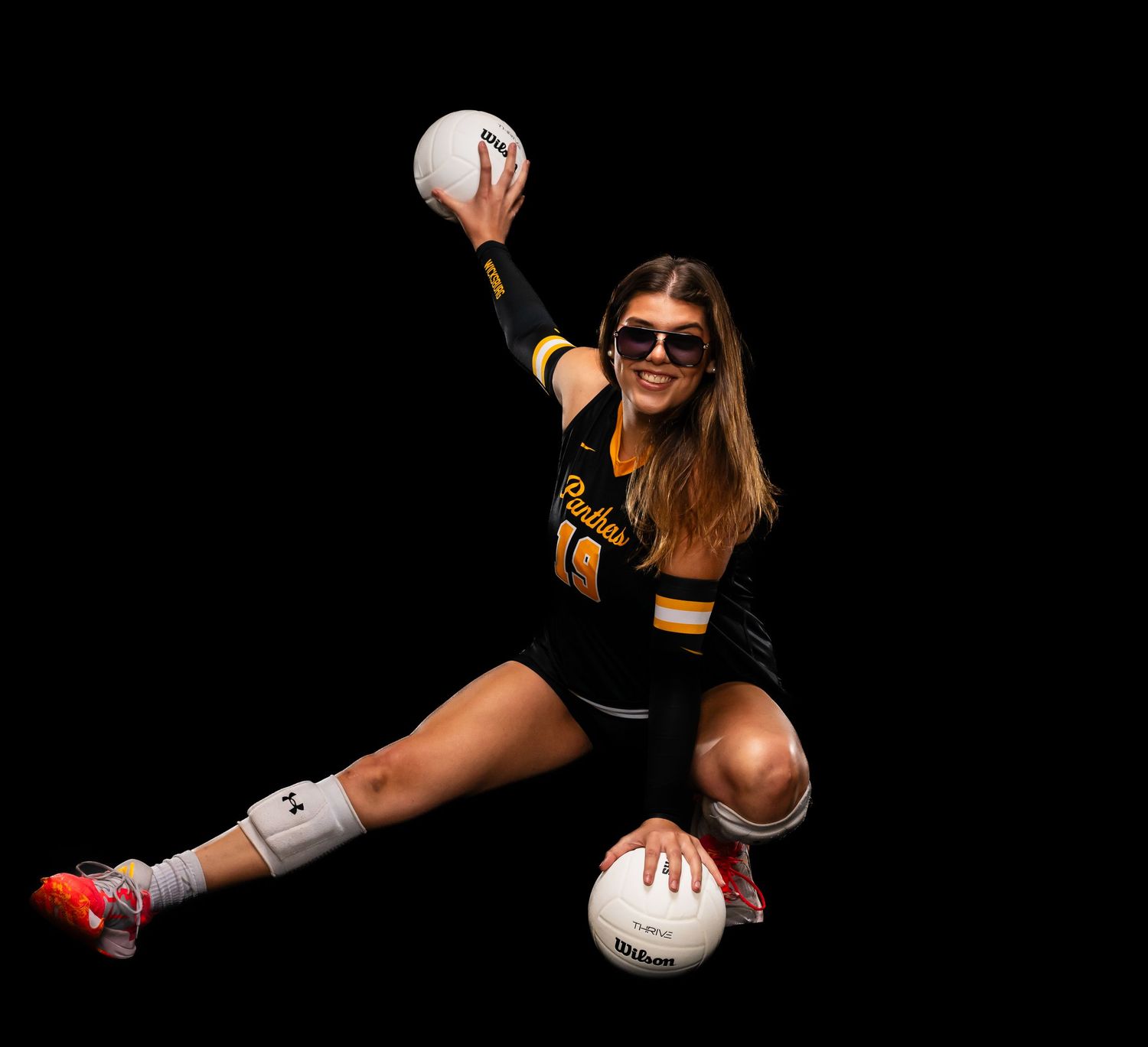 Female volleyball player posing dynamically with two volleyballs, wearing black and gold uniform and sunglasses.