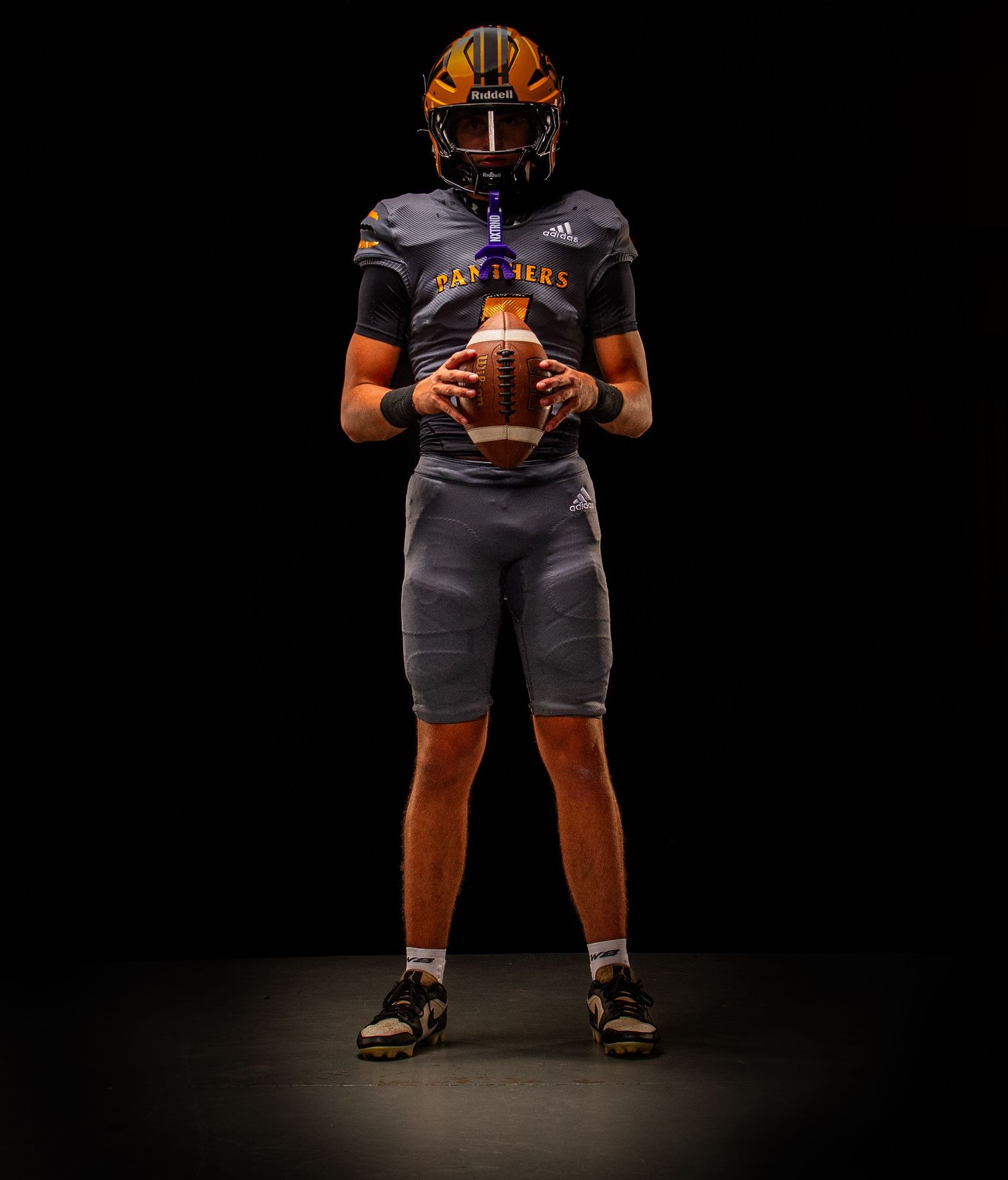 Football player in gray uniform with orange helmet holds ball, wearing Adidas gear in dramatic dark studio lighting.