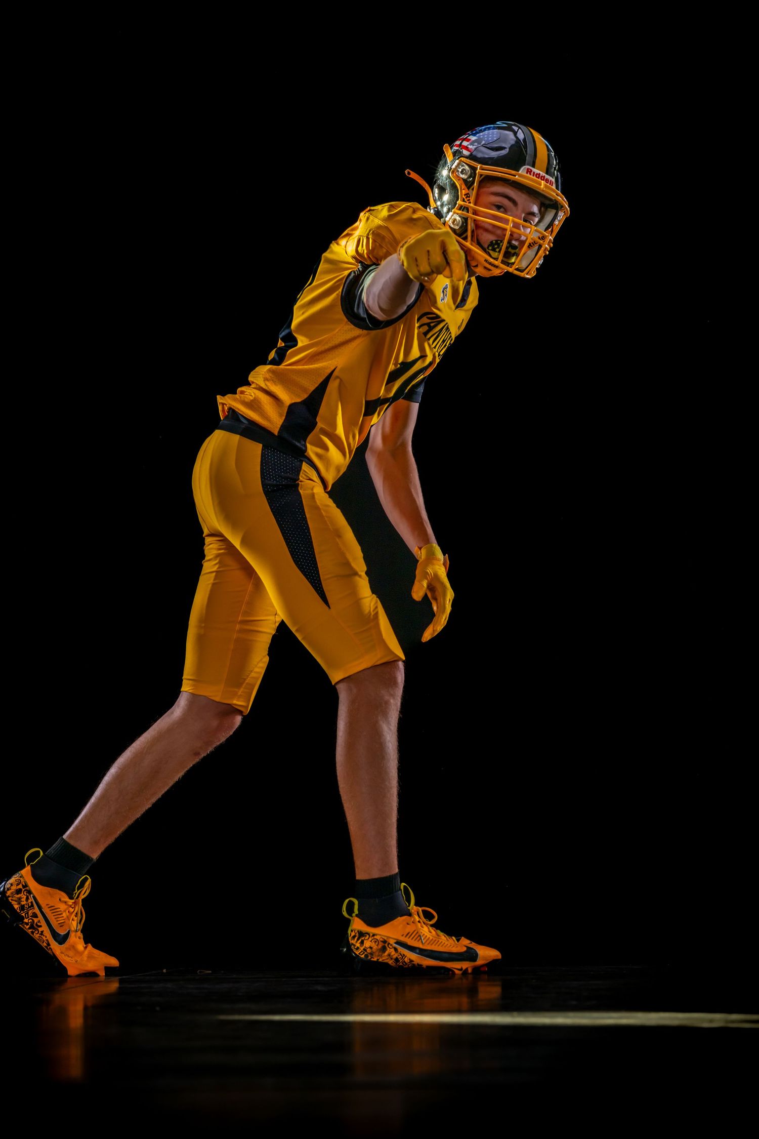 Young American football player in gold uniform crouching in ready stance against black background.