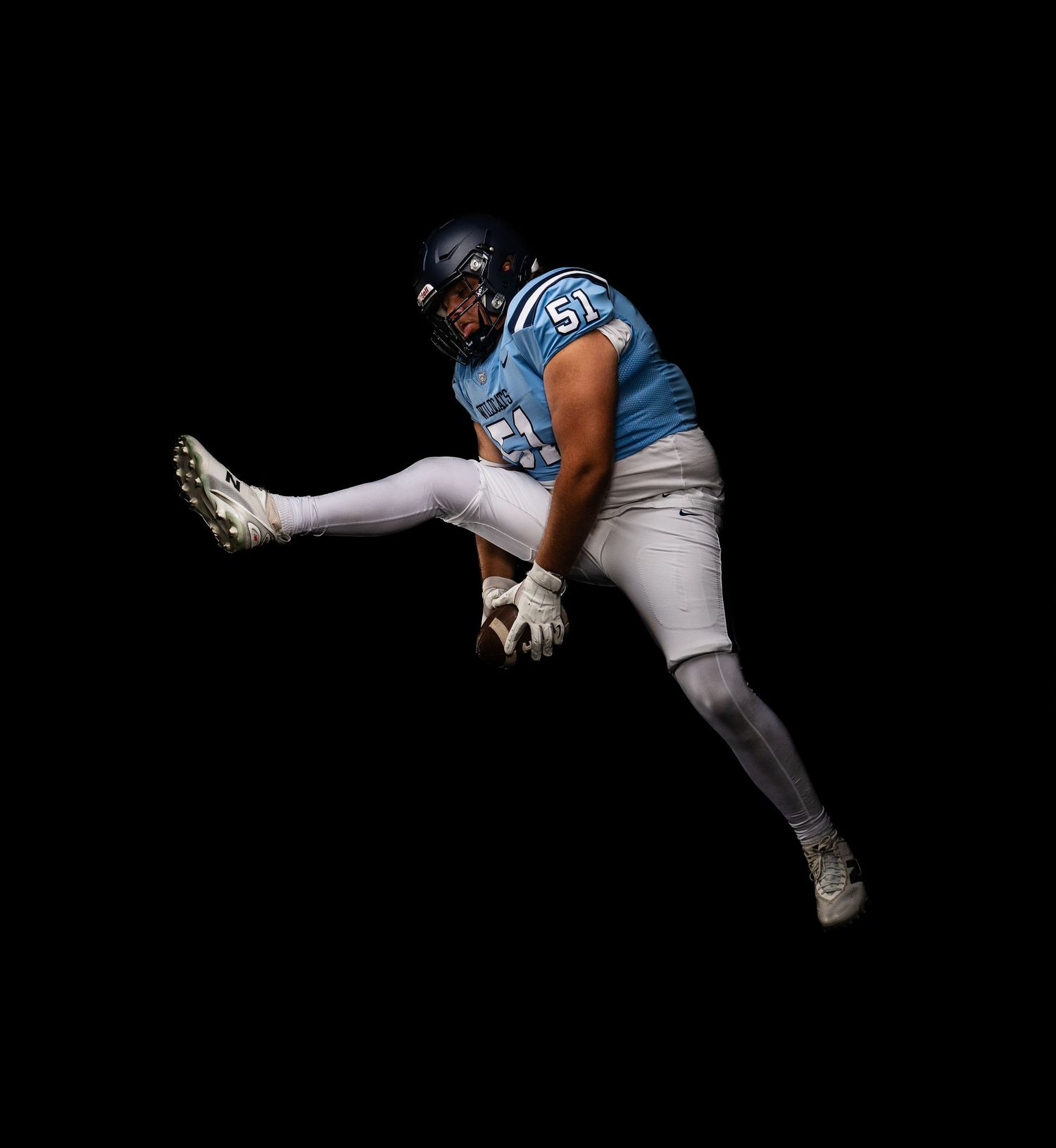 Football player #51 in light blue jersey performing a high kick against a black background.