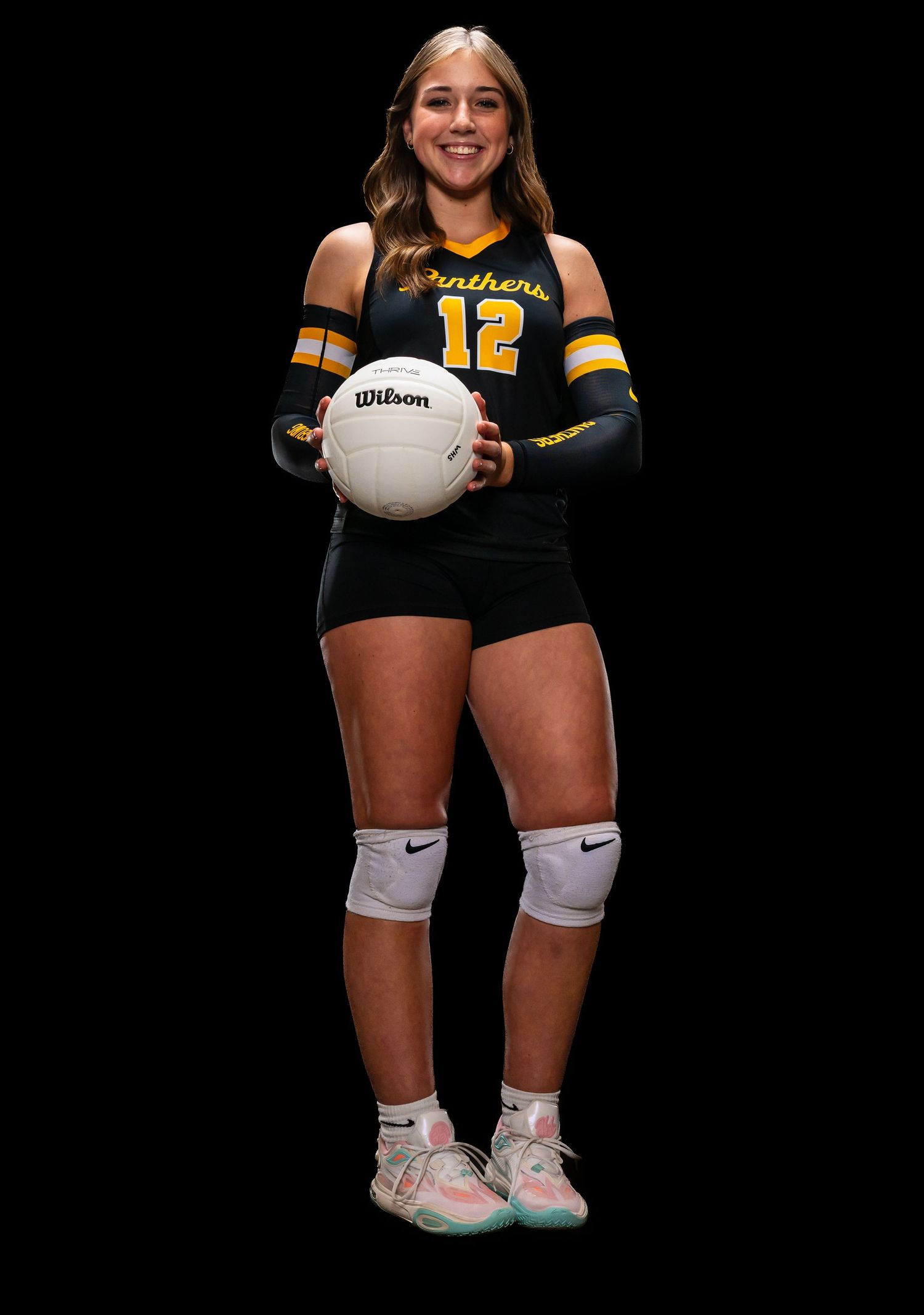 Smiling volleyball player #12 in Panthers uniform holding a Wilson ball, wearing knee pads and colorful sneakers.