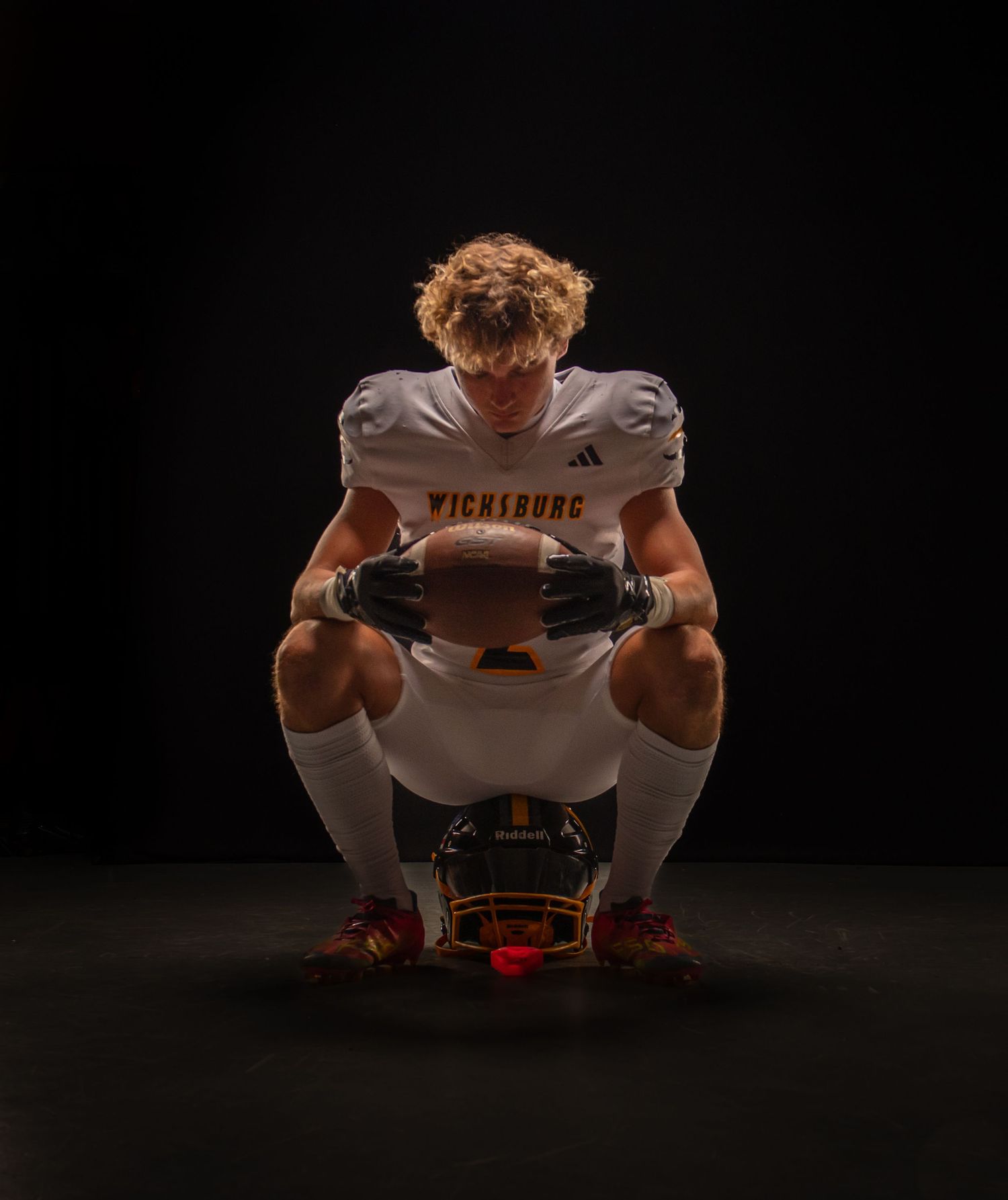 Football player in Vicksburg uniform crouching with helmet and ball in dramatic dark studio lighting.