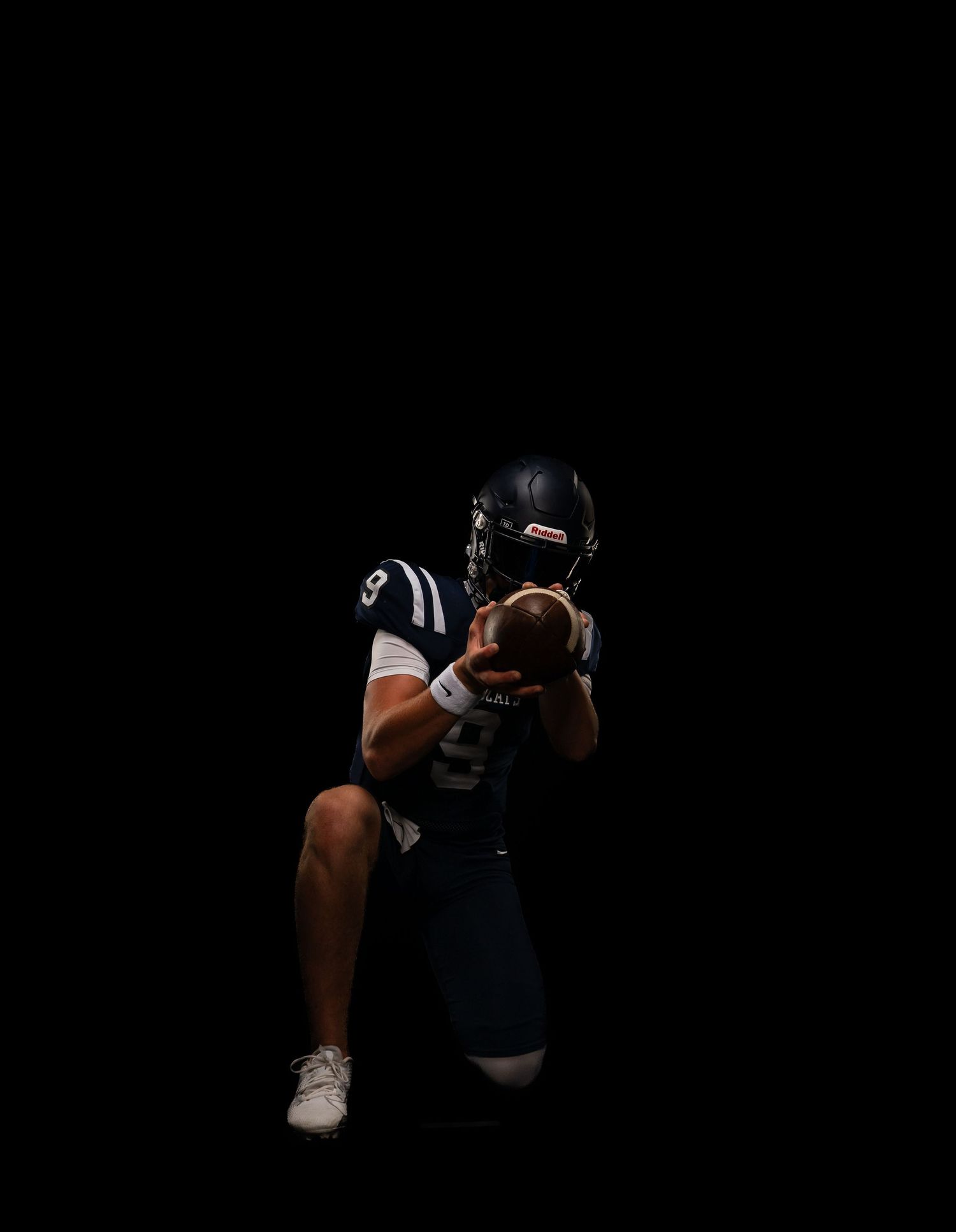 A football player in navy uniform and helmet crouches in darkness, holding a football in a dramatic studio pose.