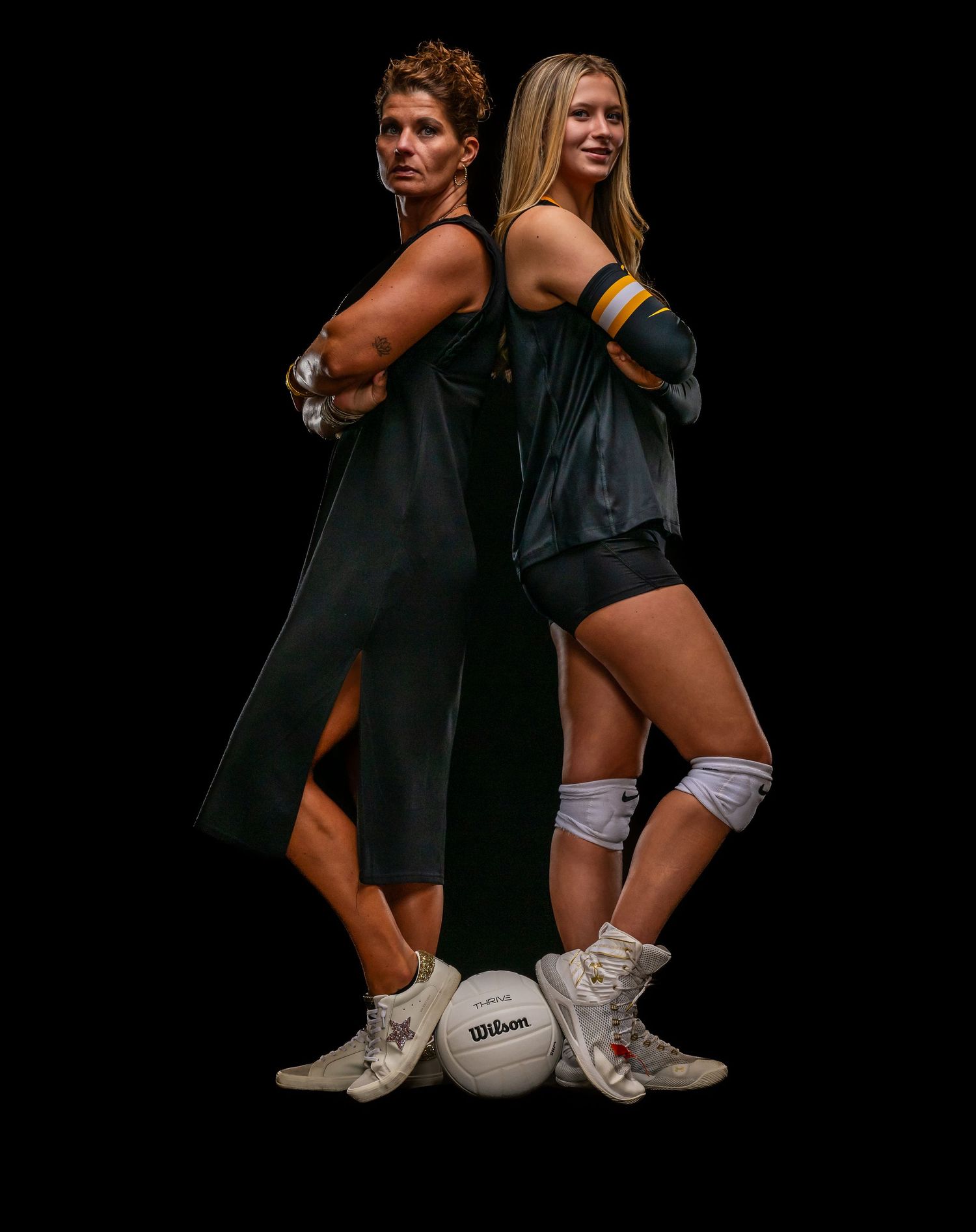 Two female volleyball players pose back-to-back with a Wilson volleyball between them on a dark studio background.