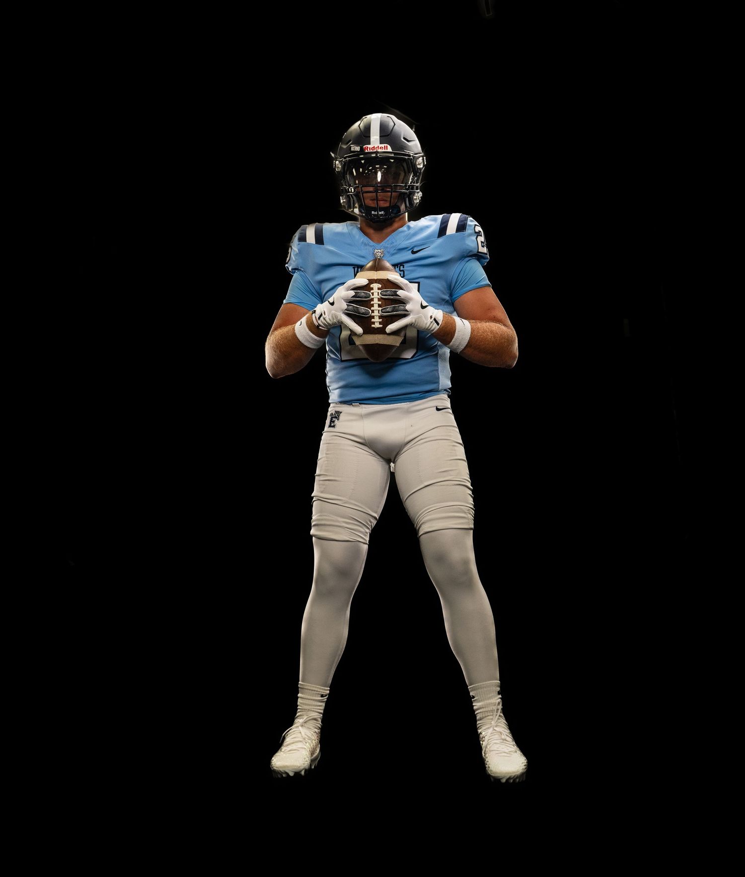 Football player in light blue jersey and white pants holds a football, wearing a black helmet against a dark background.