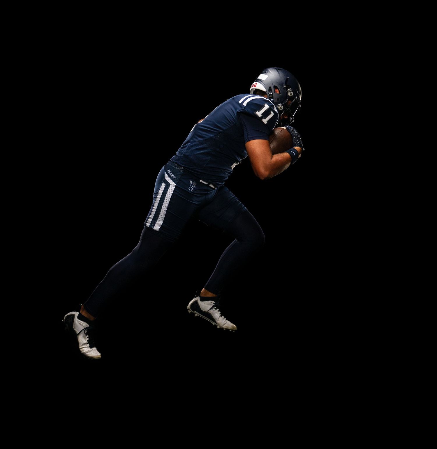 Football player in navy uniform and helmet catches ball mid-run against a dramatic black background.