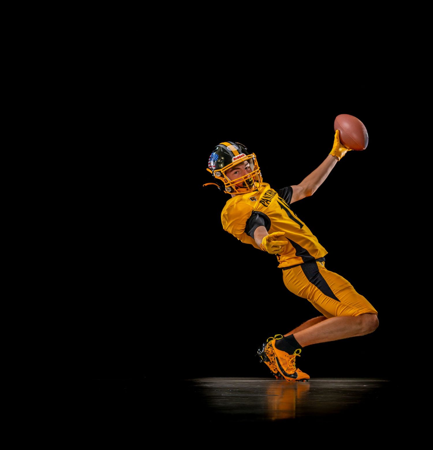 Football player in yellow uniform leaping dramatically to catch ball against black background.