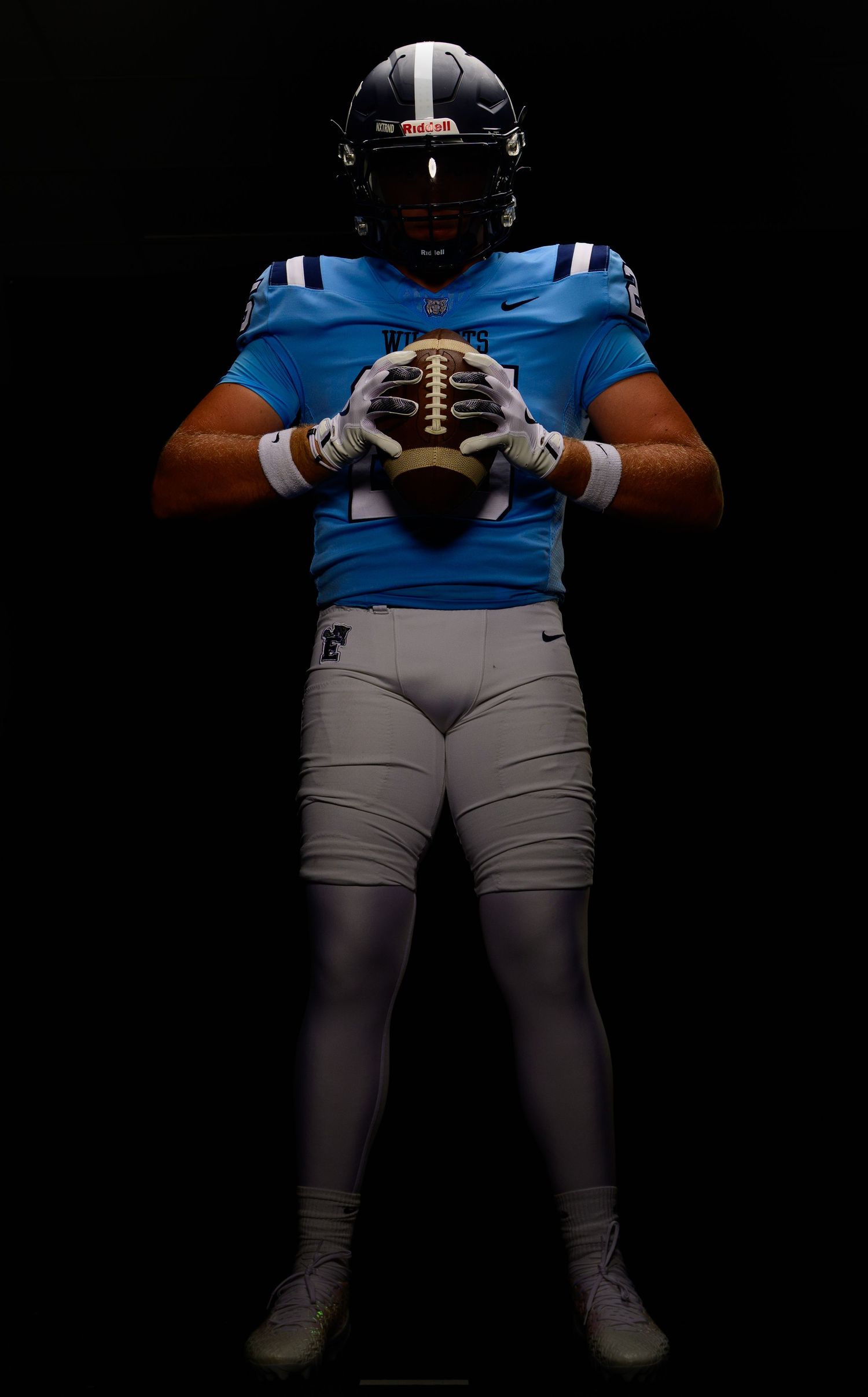 Football player in light blue jersey and gray pants holds a football, posing in dramatic dark studio lighting.