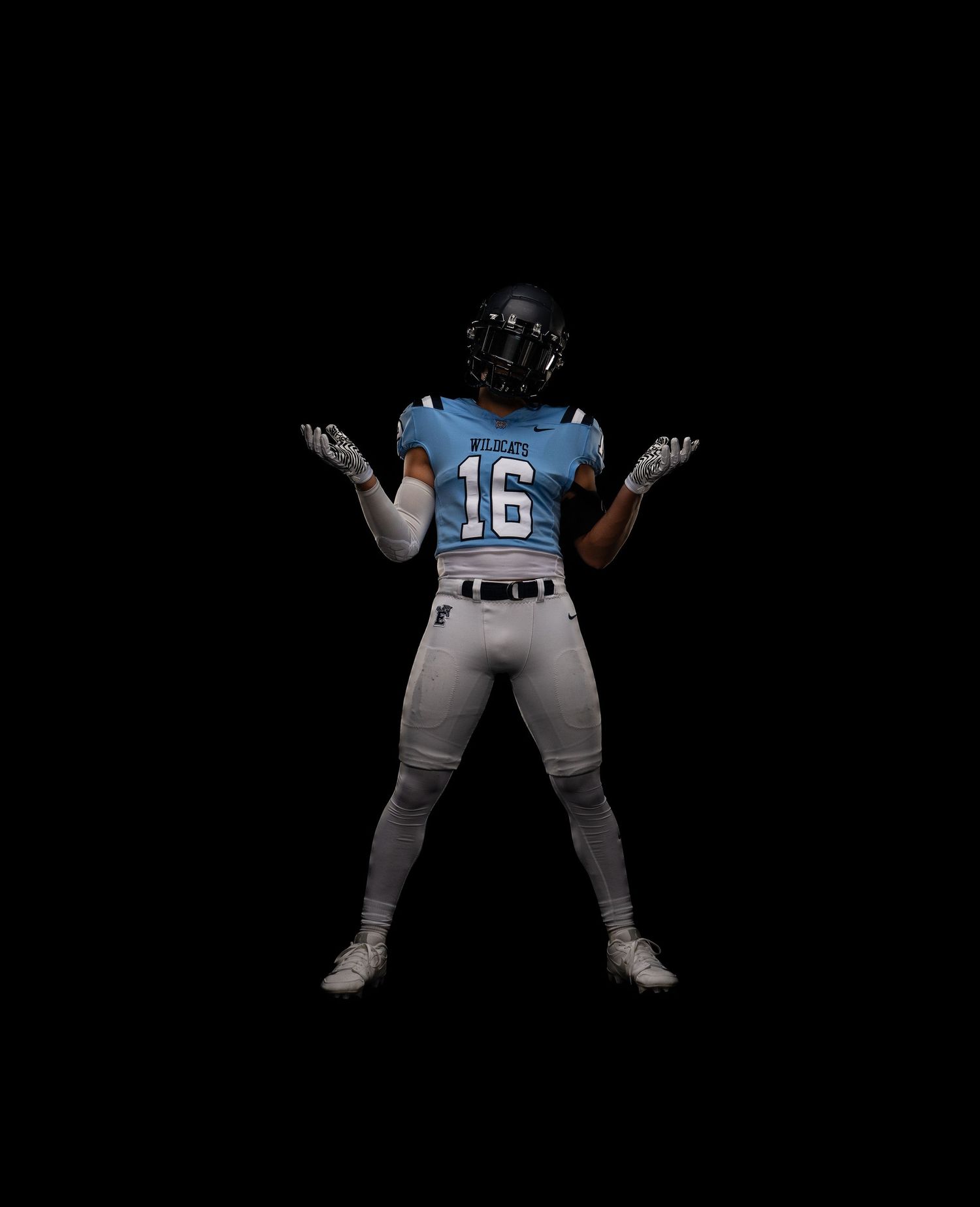 Football player wearing number 16 light blue jersey stands with arms raised against black background.