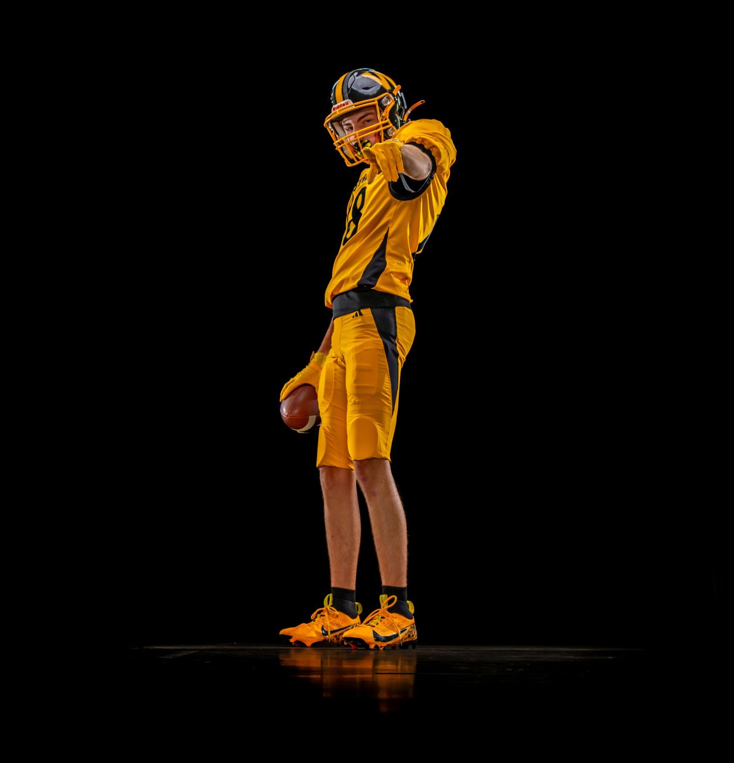 Youth football player in gold uniform throws ball in side profile pose against black background.