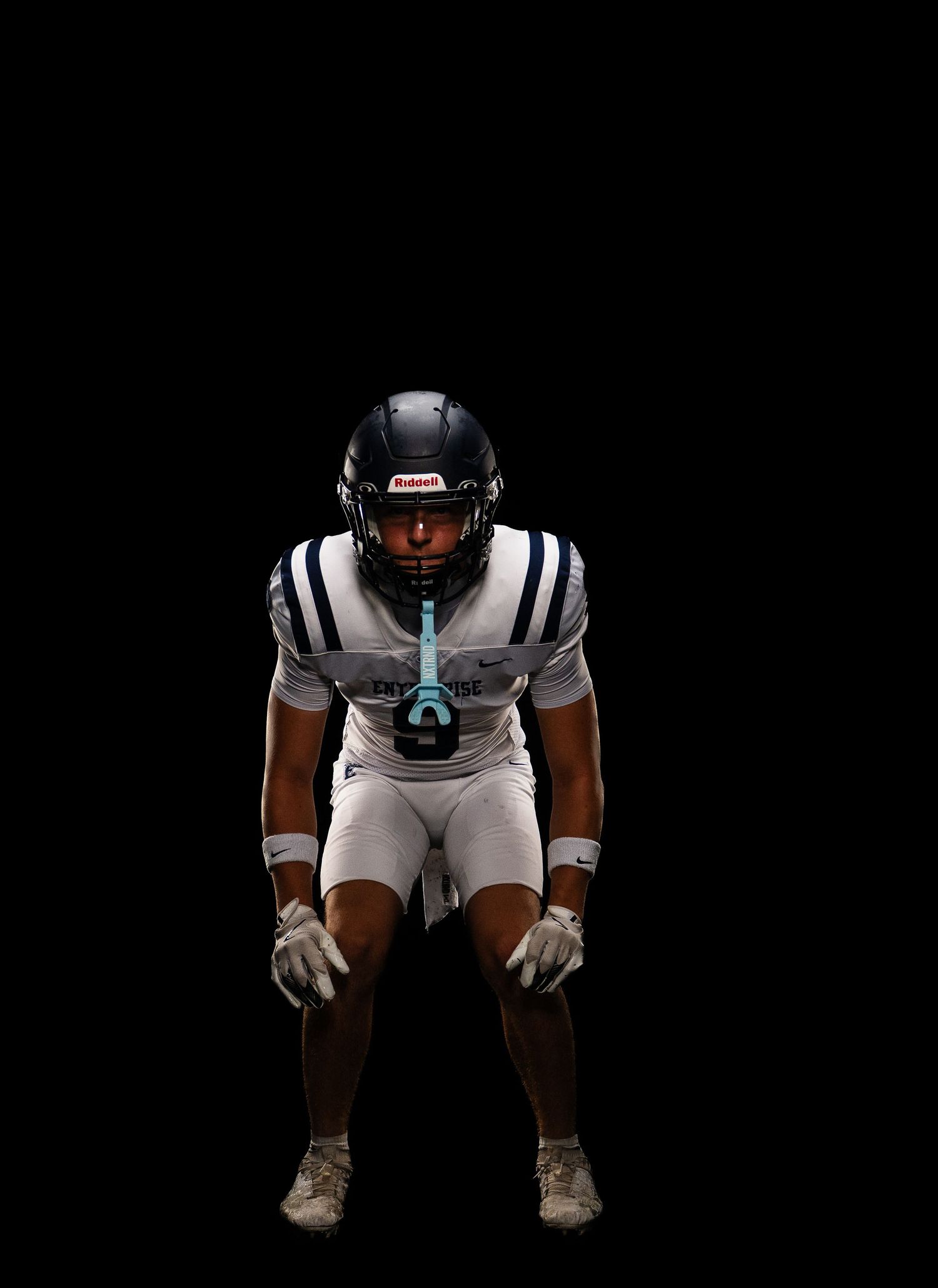 Football player in white uniform crouches in ready stance against black background.