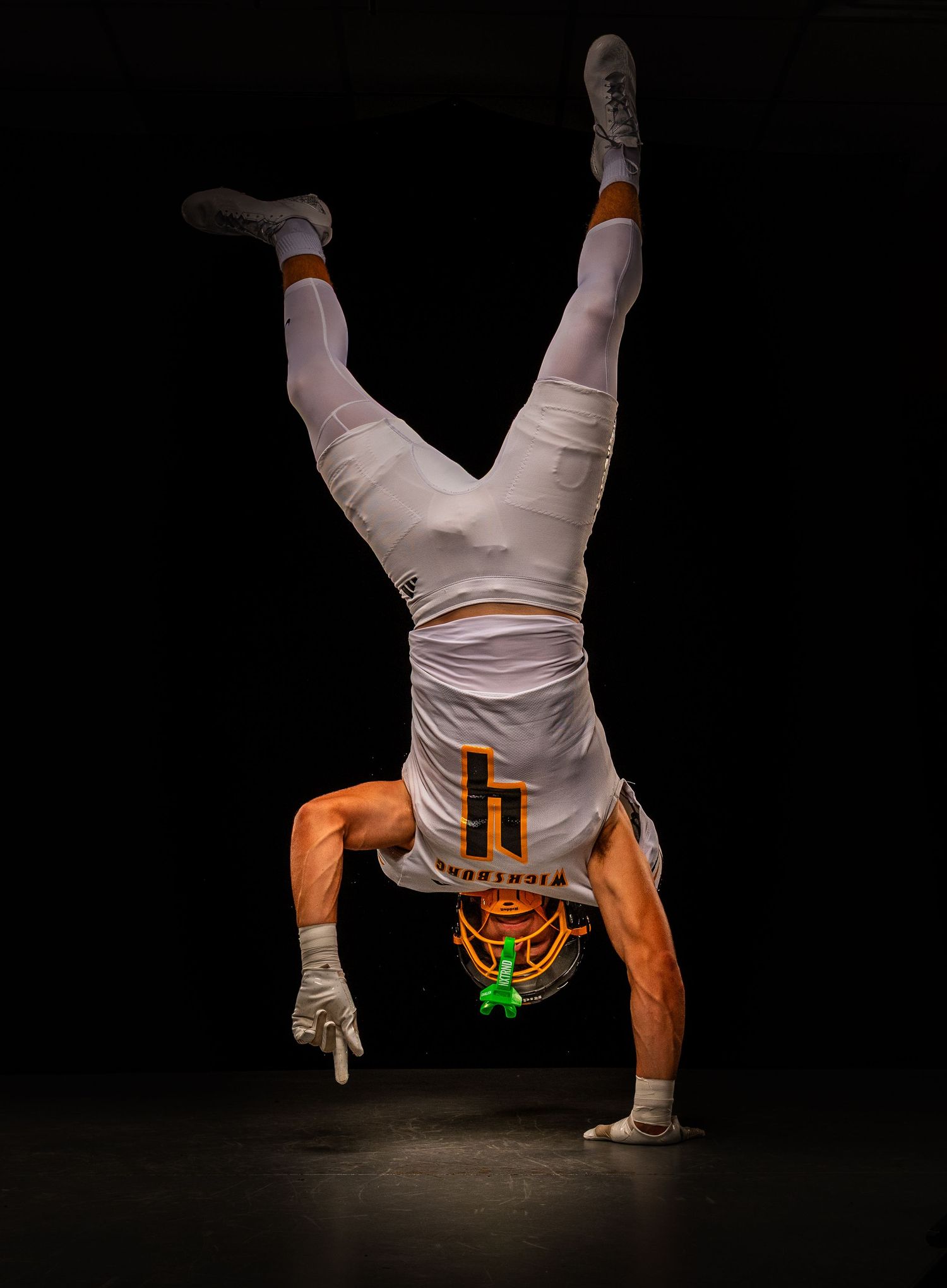Athlete in orange and white uniform performs handstand against dark backdrop.