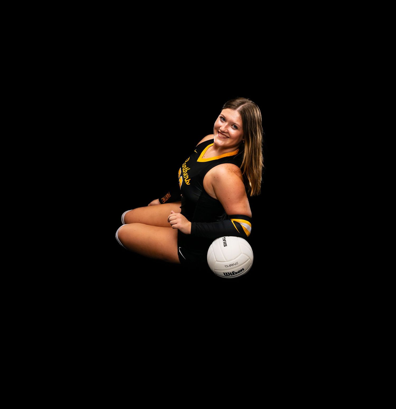 Smiling female volleyball player in black and gold Panthers uniform sitting on floor with volleyball.