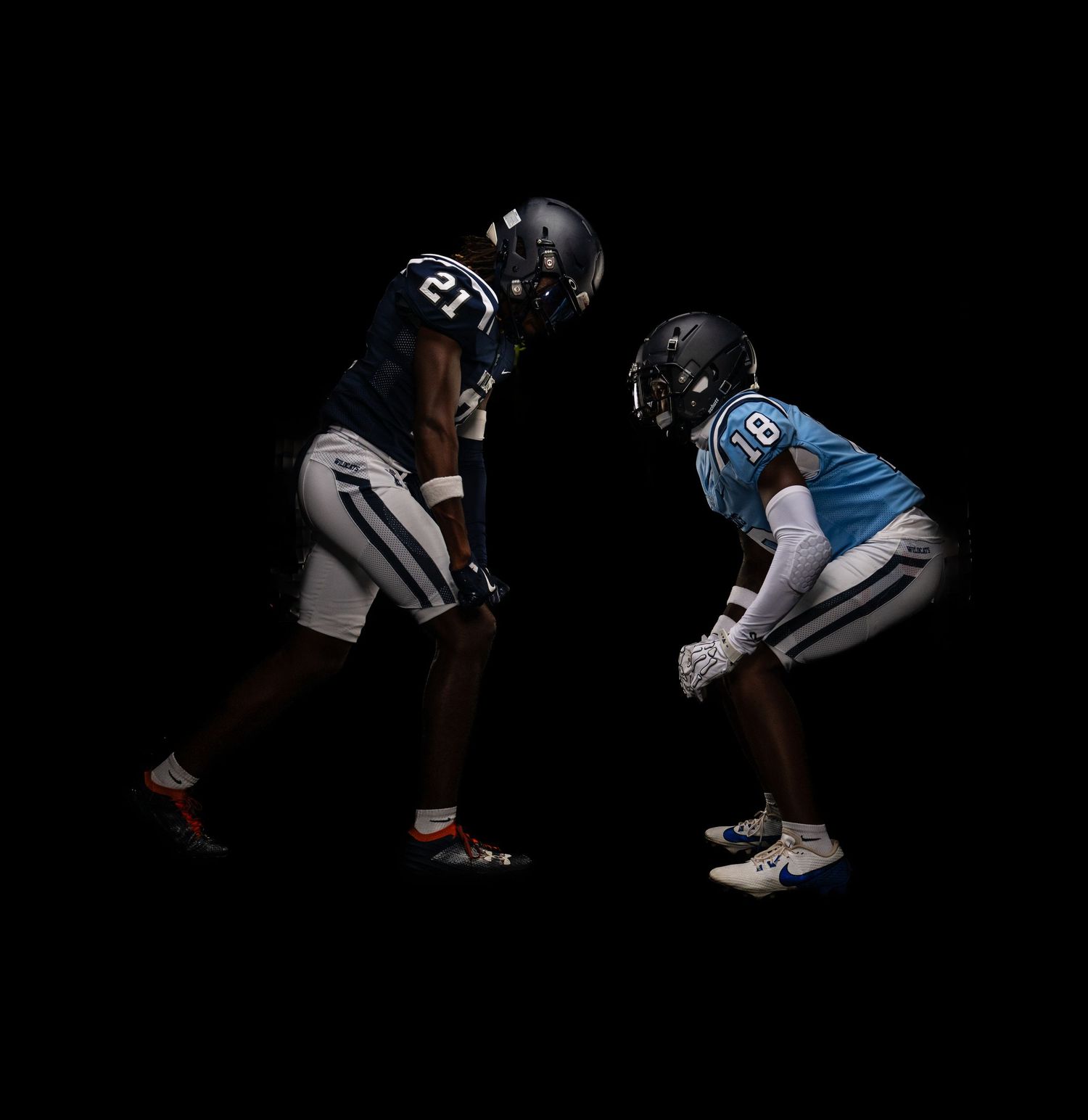 Two American football players in opposing uniforms face off in a dramatic dark studio setting.