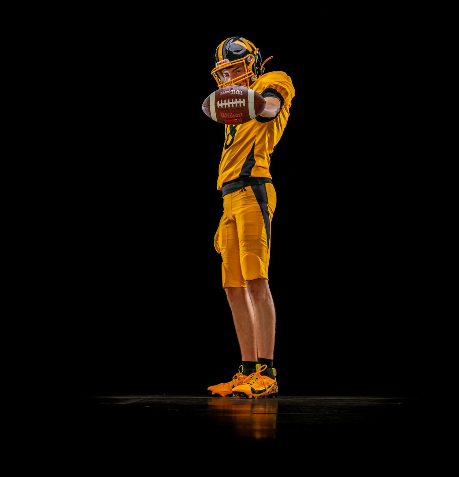 Football player in gold uniform poised to throw a football against a dark background.