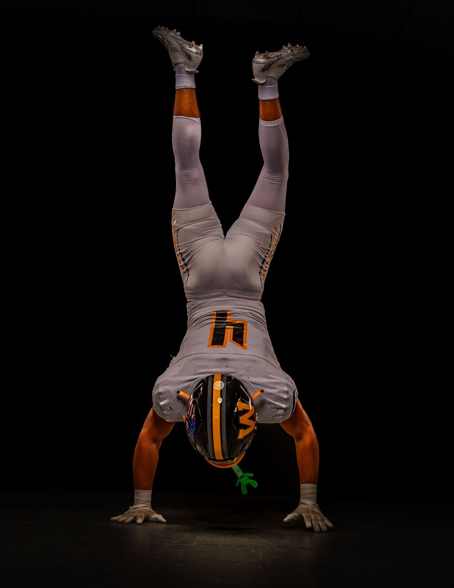 Football player in gray and orange uniform doing a handstand on dark background.