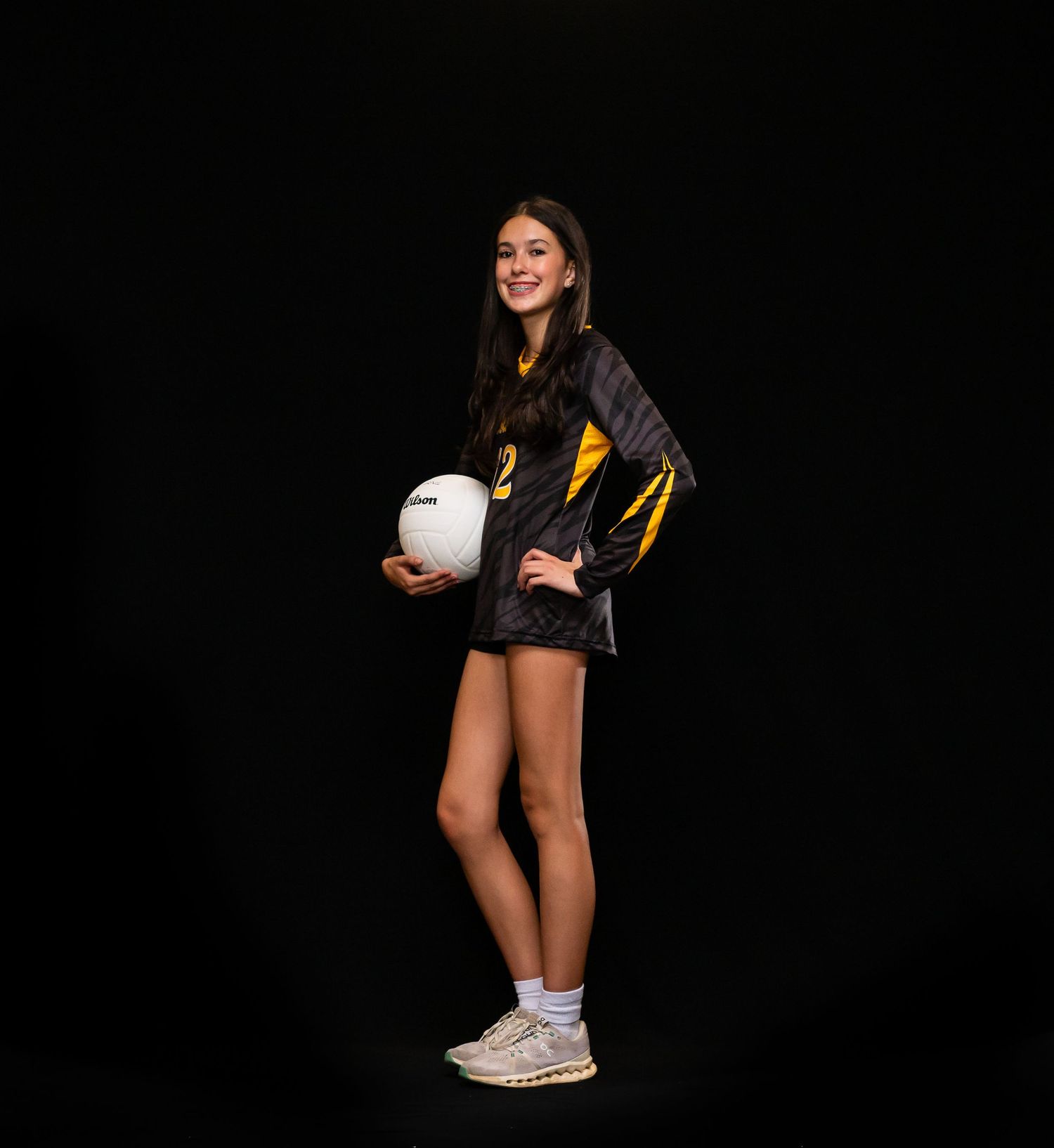Tall female volleyball player in black and gold uniform holding volleyball against dark background.
