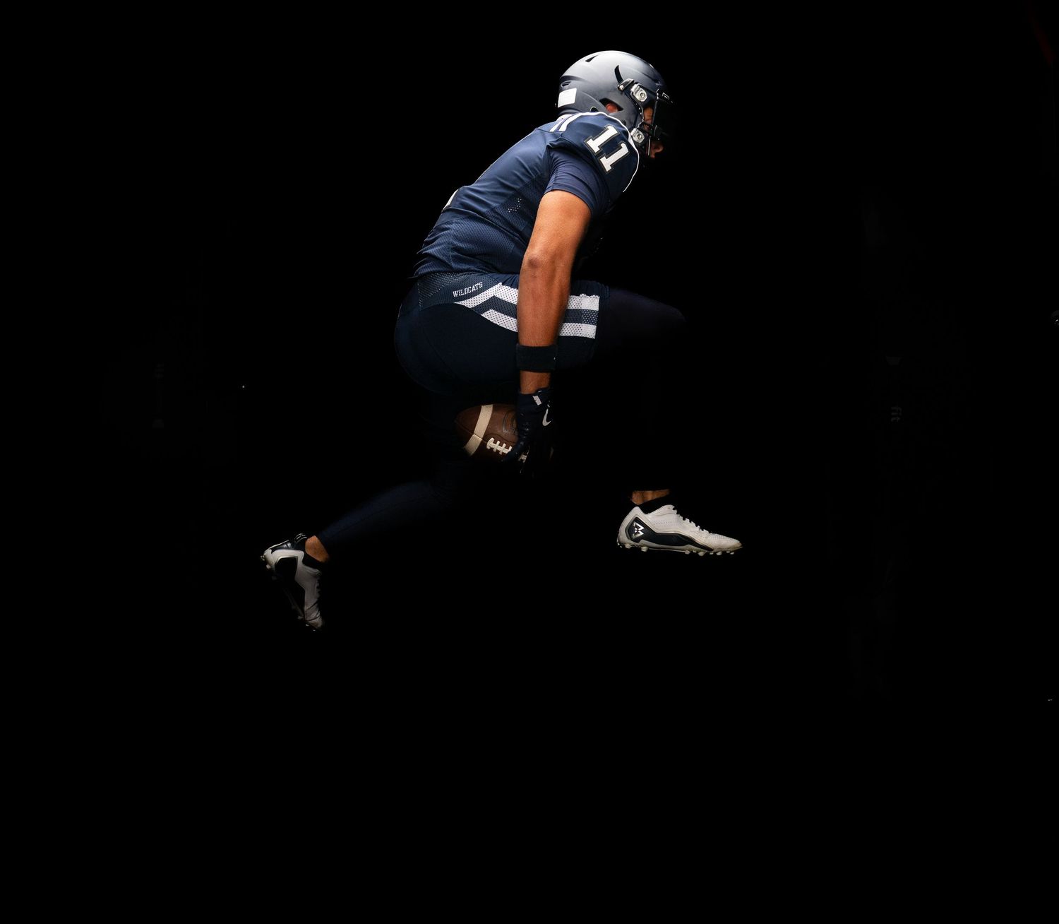 Football player in navy blue uniform and white cleats leaping dynamically against a dark background.