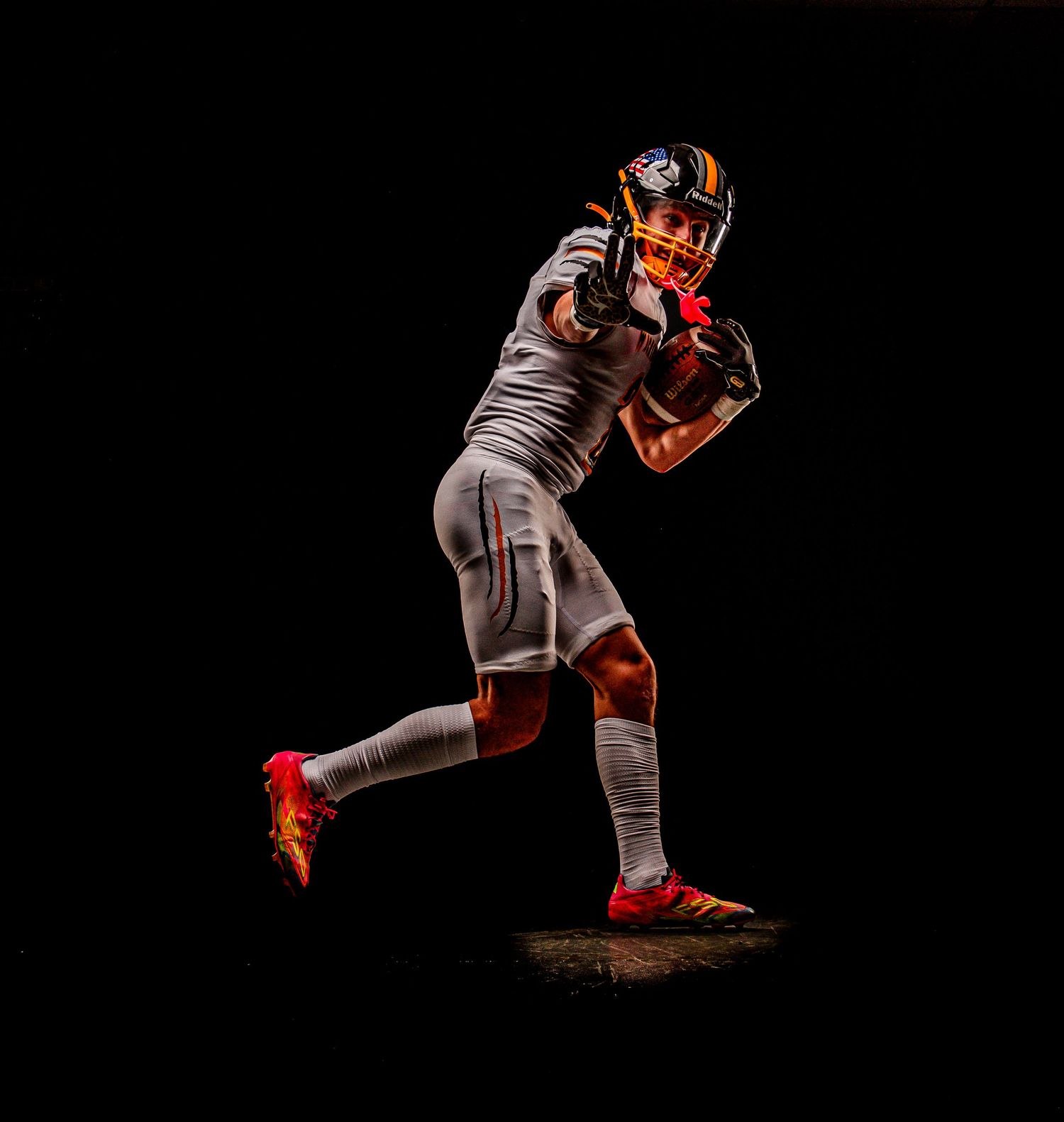 Young football player in white uniform catches ball mid-run against dramatic black background with vibrant lighting.