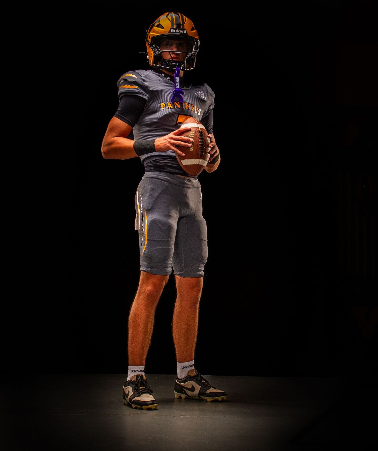 Football player in protective gear holding a football against black background.