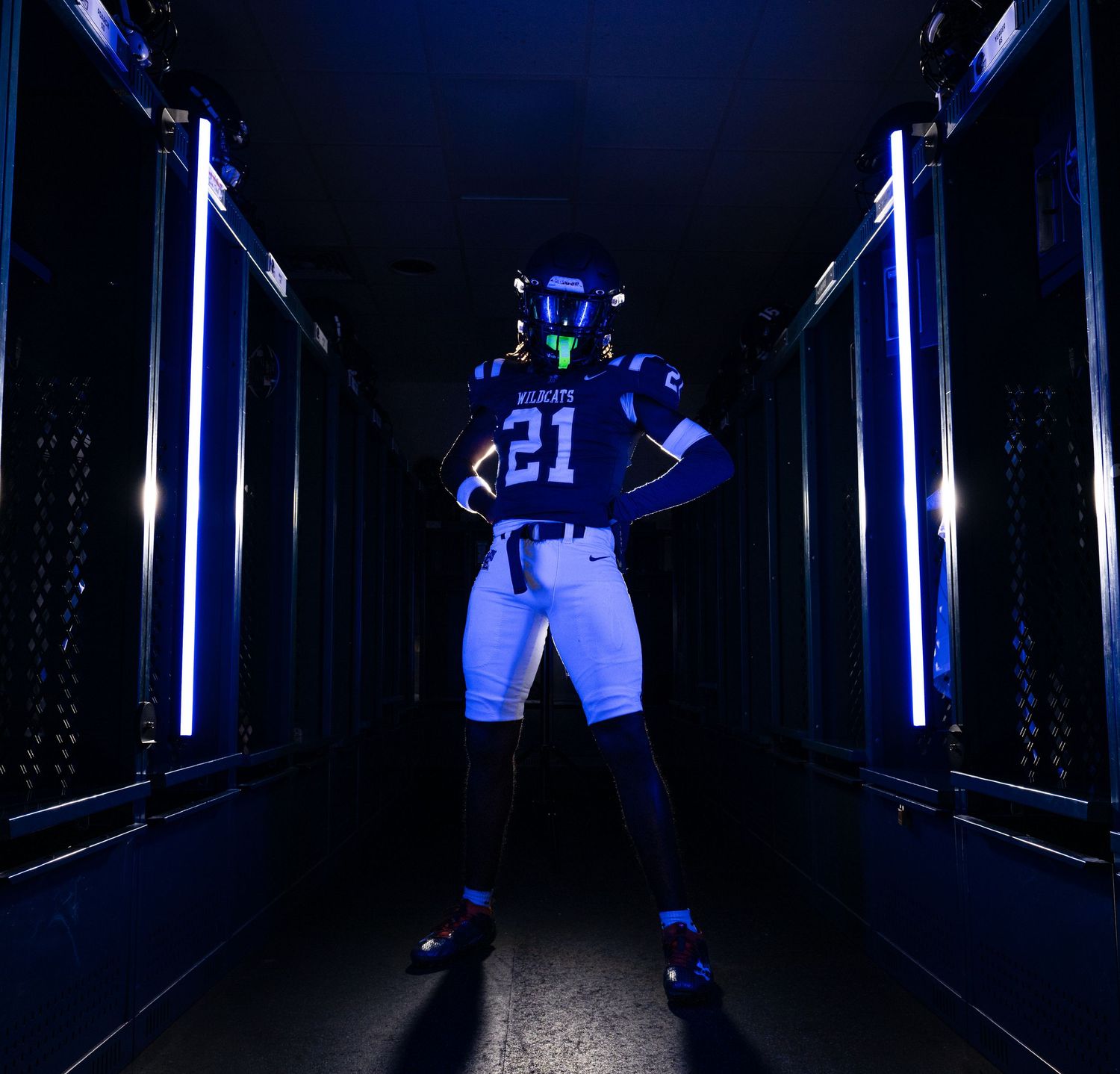 Football player wearing number 21 jersey poses confidently in a dark locker room corridor with blue LED lighting.