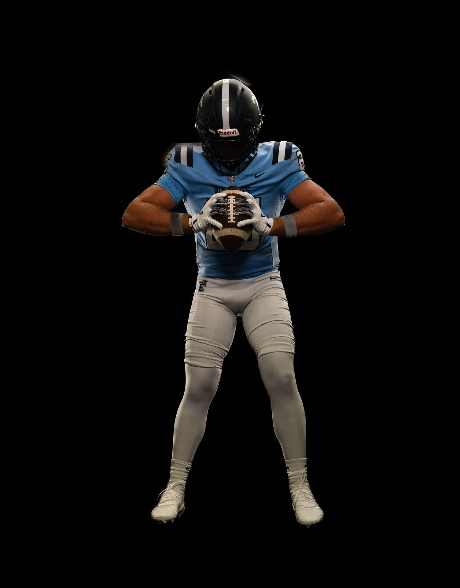 Football player in blue jersey and white pants holding a football, wearing a black helmet on dark background.