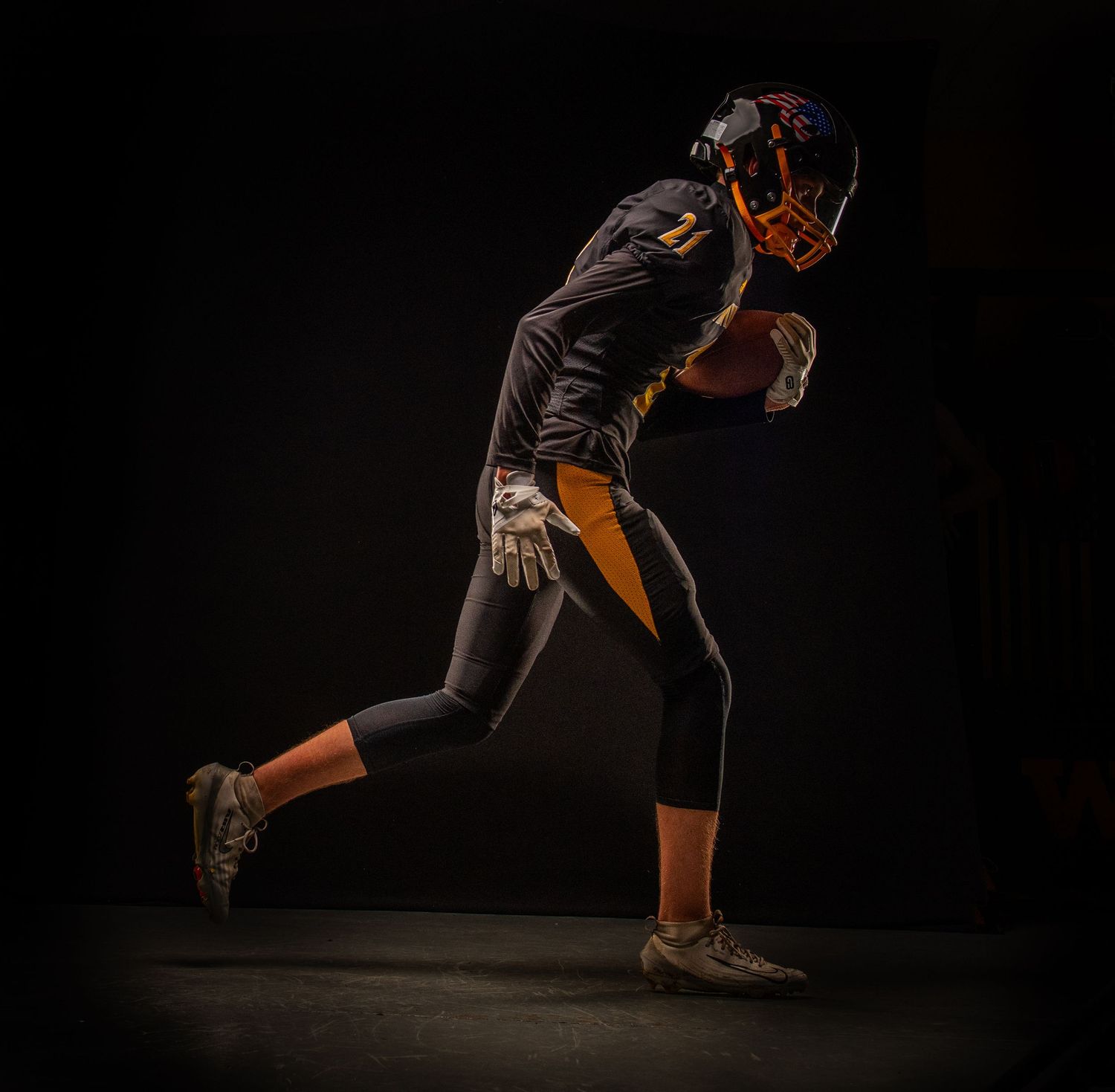 Football player in black and gold uniform running with ball against dark background.