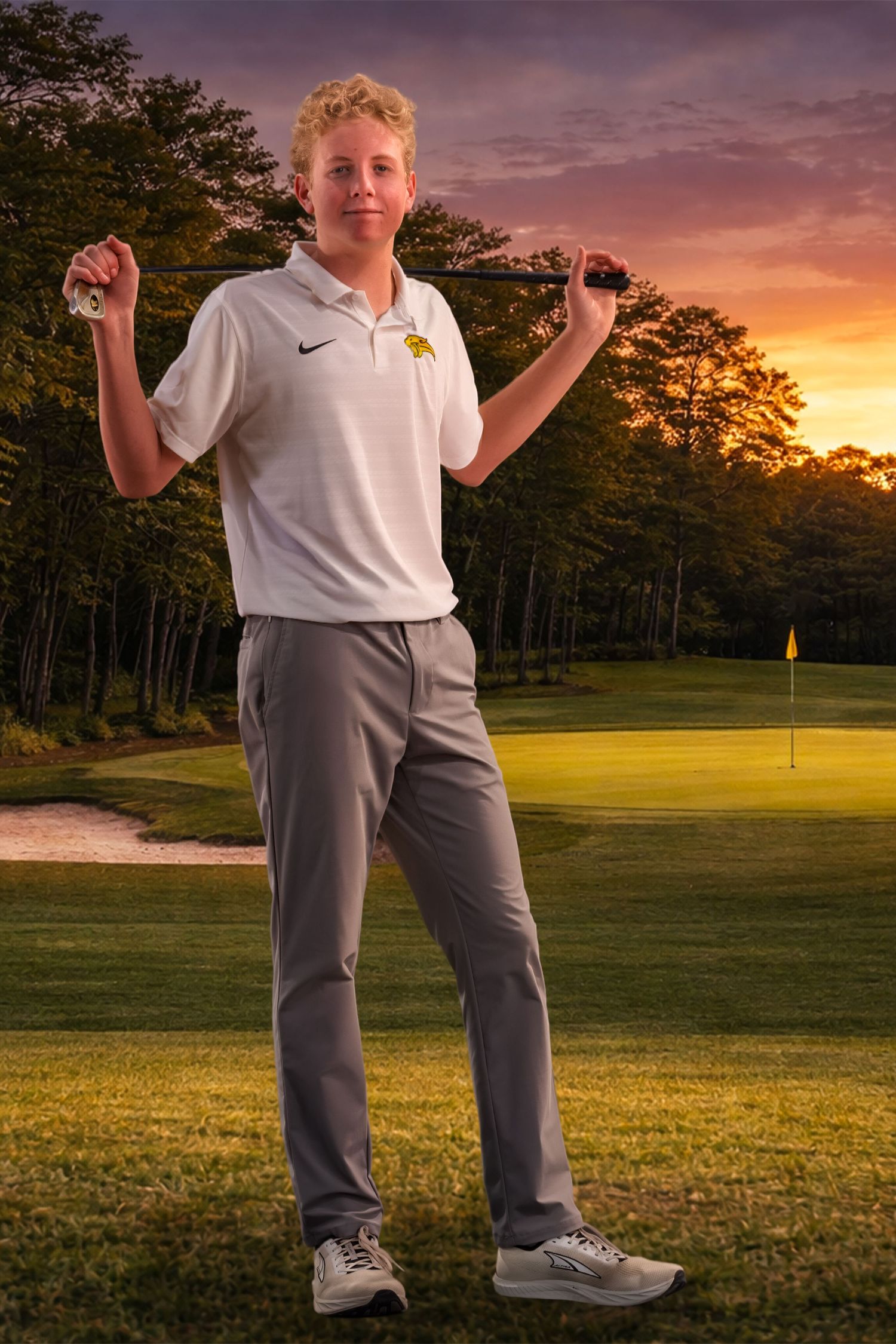 Golfer in white polo and grey pants demonstrates stance on golf course at sunset.