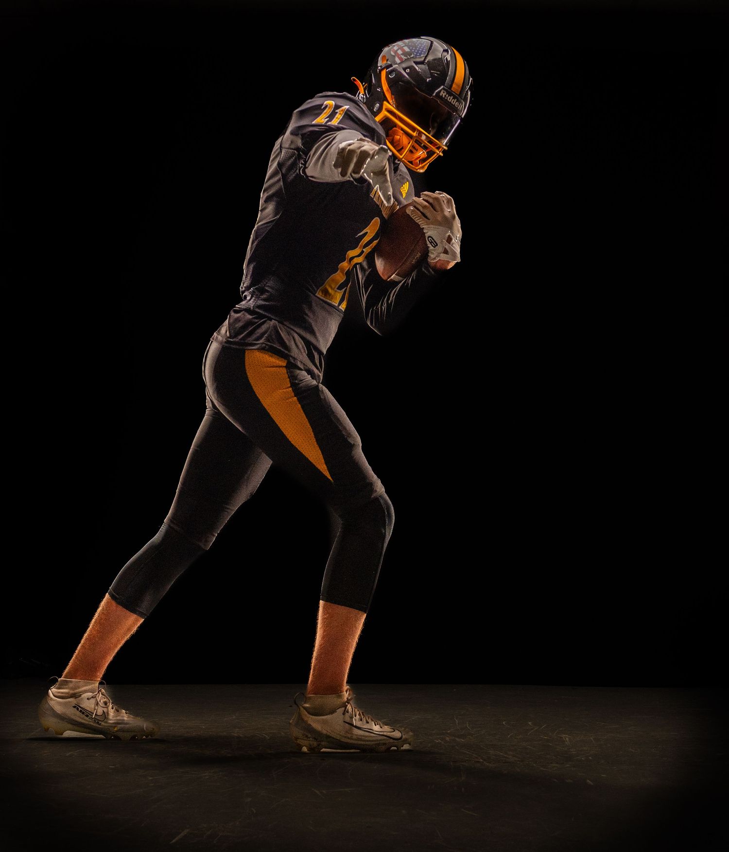 American football player in black and gold uniform holding ball in dynamic pose against dark background.