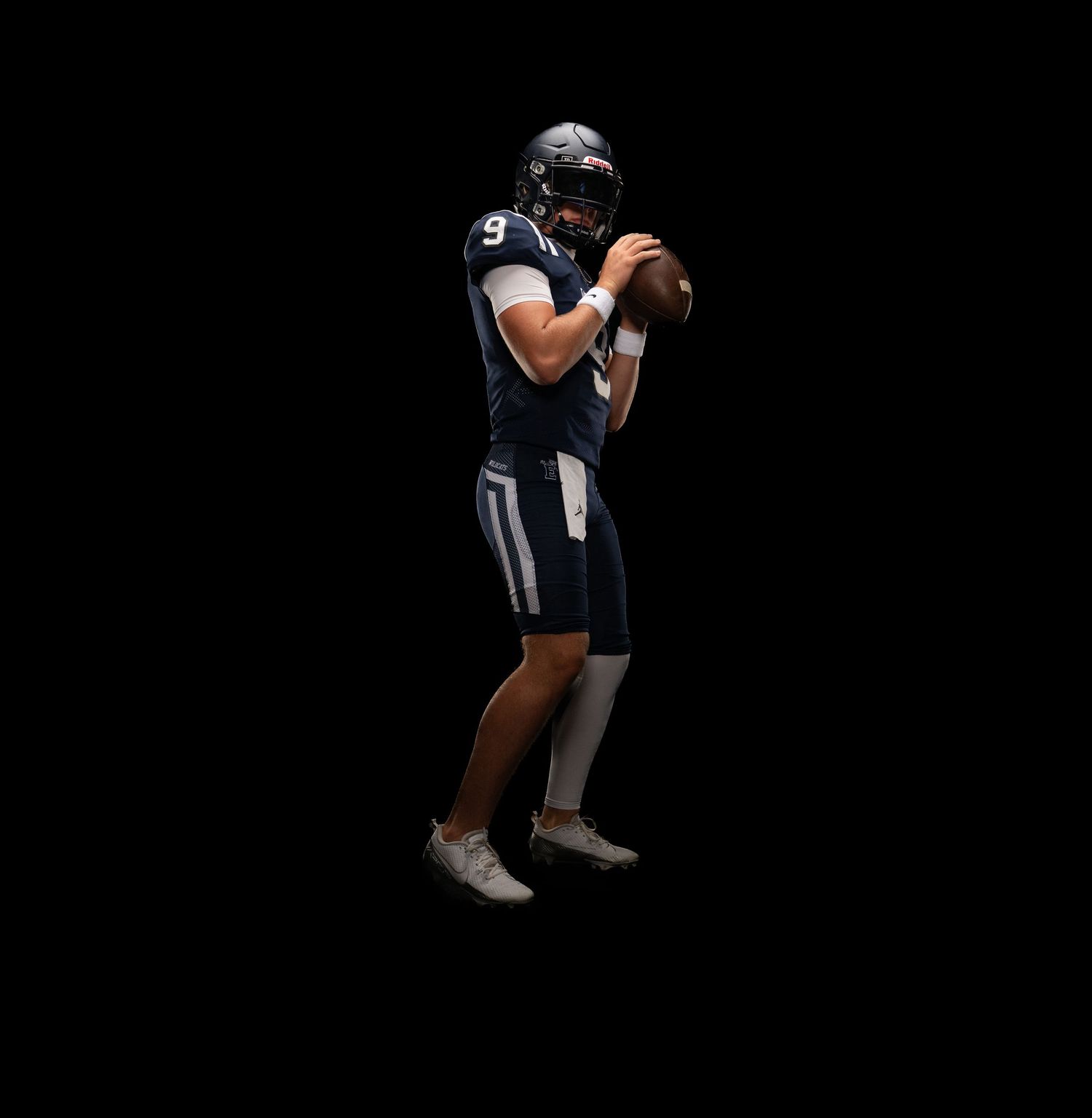 Football player wearing navy blue jersey number 9 holds ball in throwing stance against black background.